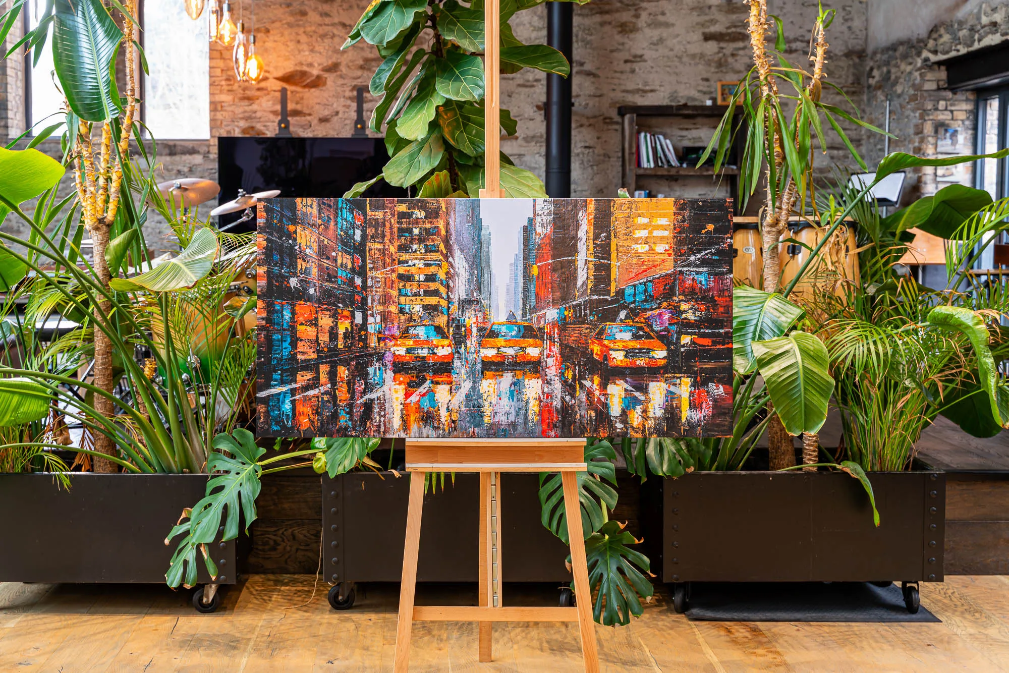 Downtown Reflections, a limited edition print by Paul Kenton, on an easel among tropical plants in a brick-walled room; a textured city street of yellow taxis glows in warm orange and blue.