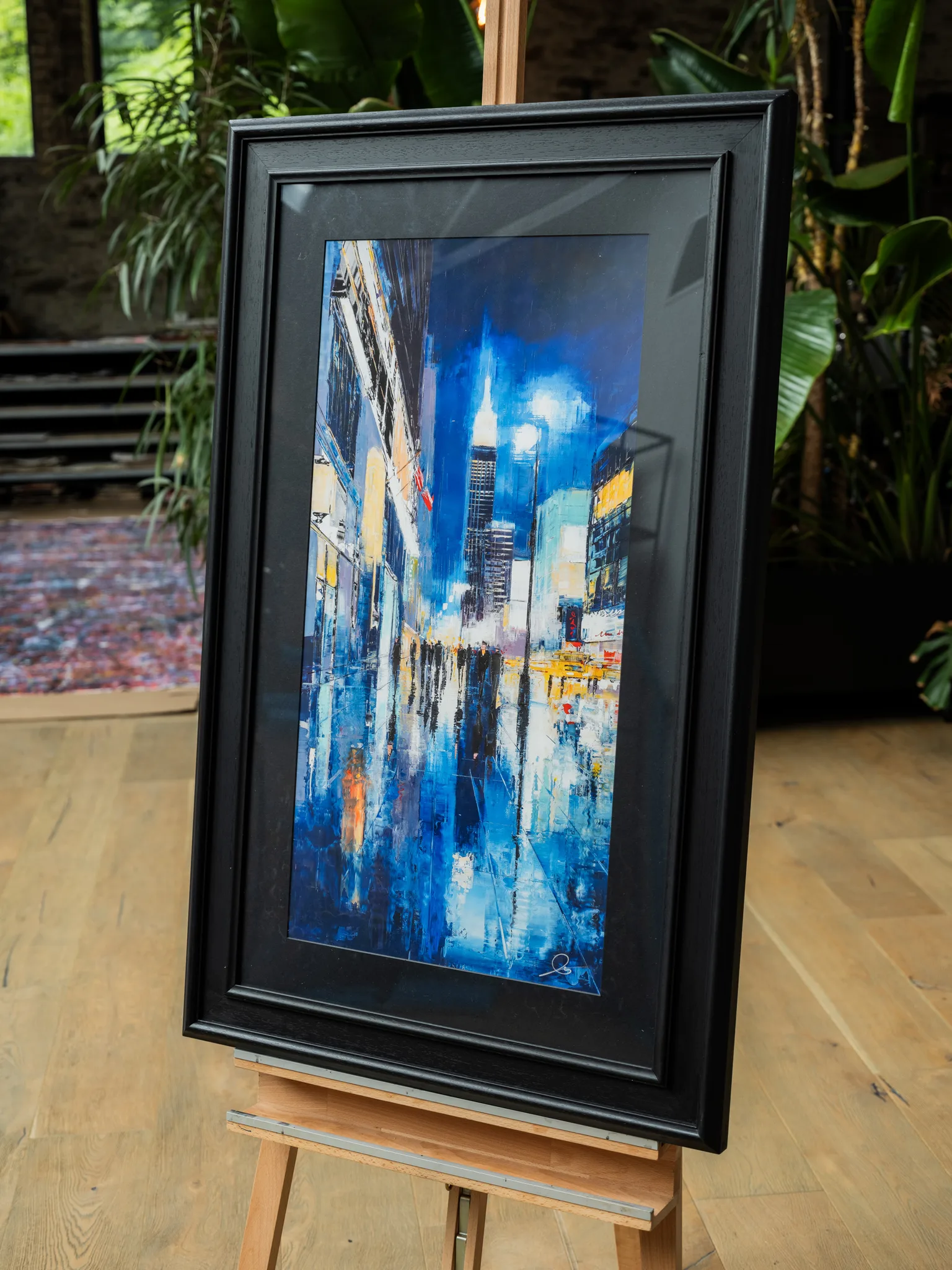 Electric City, a limited edition giclée print of New York, shown in a black frame on a wooden easel. A nocturnal street scene of tall illuminated towers and blurred blue reflections, with warm yellow lights and a lone figure at left.