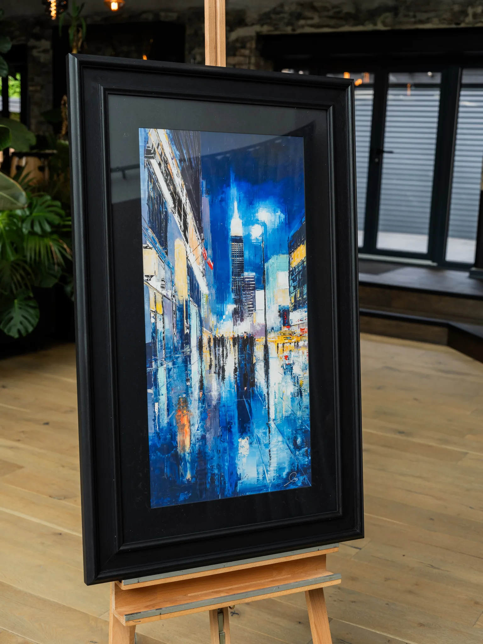 Electric City, a limited edition giclée print of New York at night, shown in a black frame on a wooden easel. A tall skyscraper glows under deep blue skies, with reflective wet streets, blurred figures and warm amber lights.