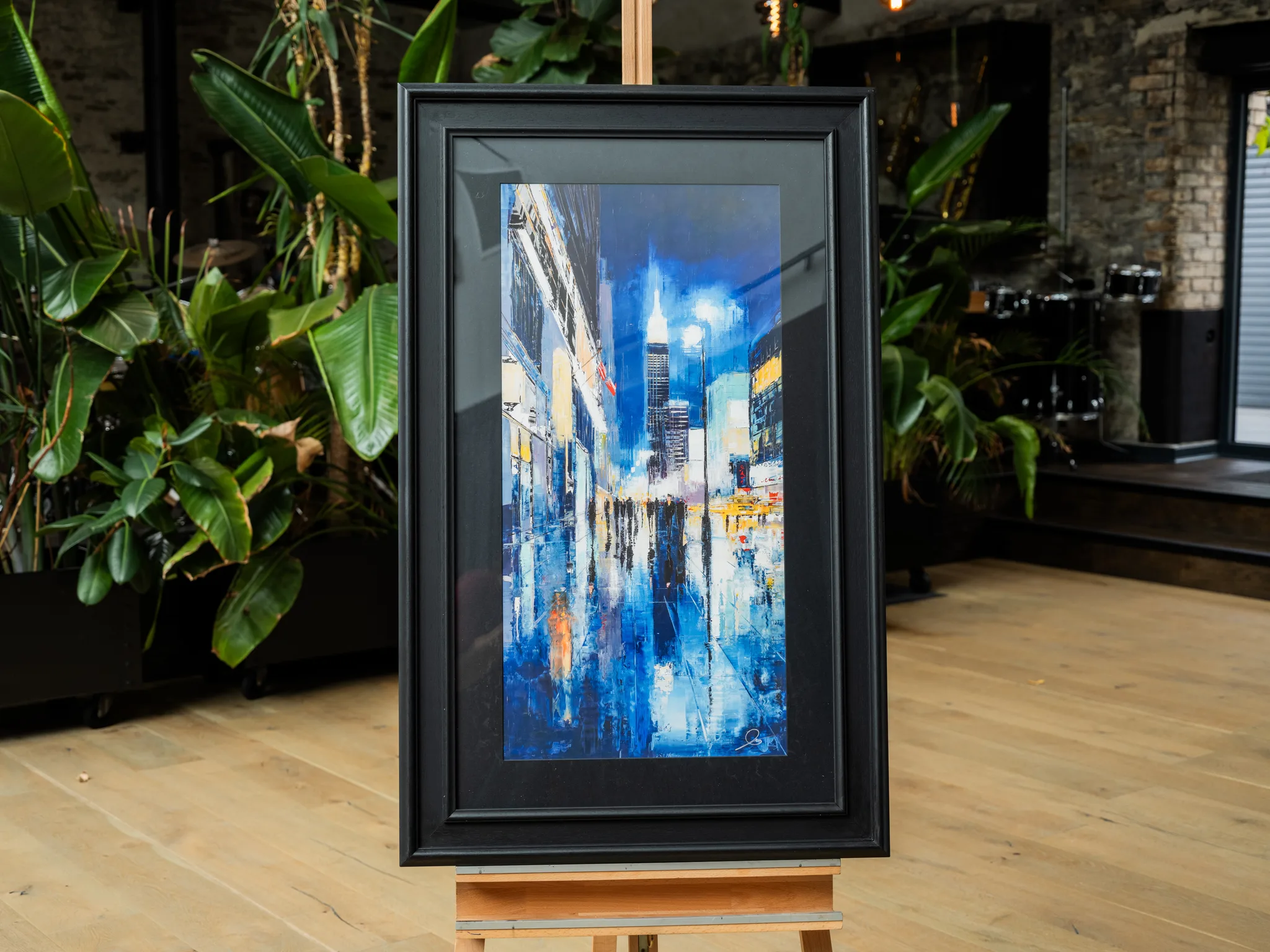 Electric City, a framed limited edition giclée print on an easel, showing a night street scene in New York with glowing towers, blue reflections on wet pavement and warm lights.