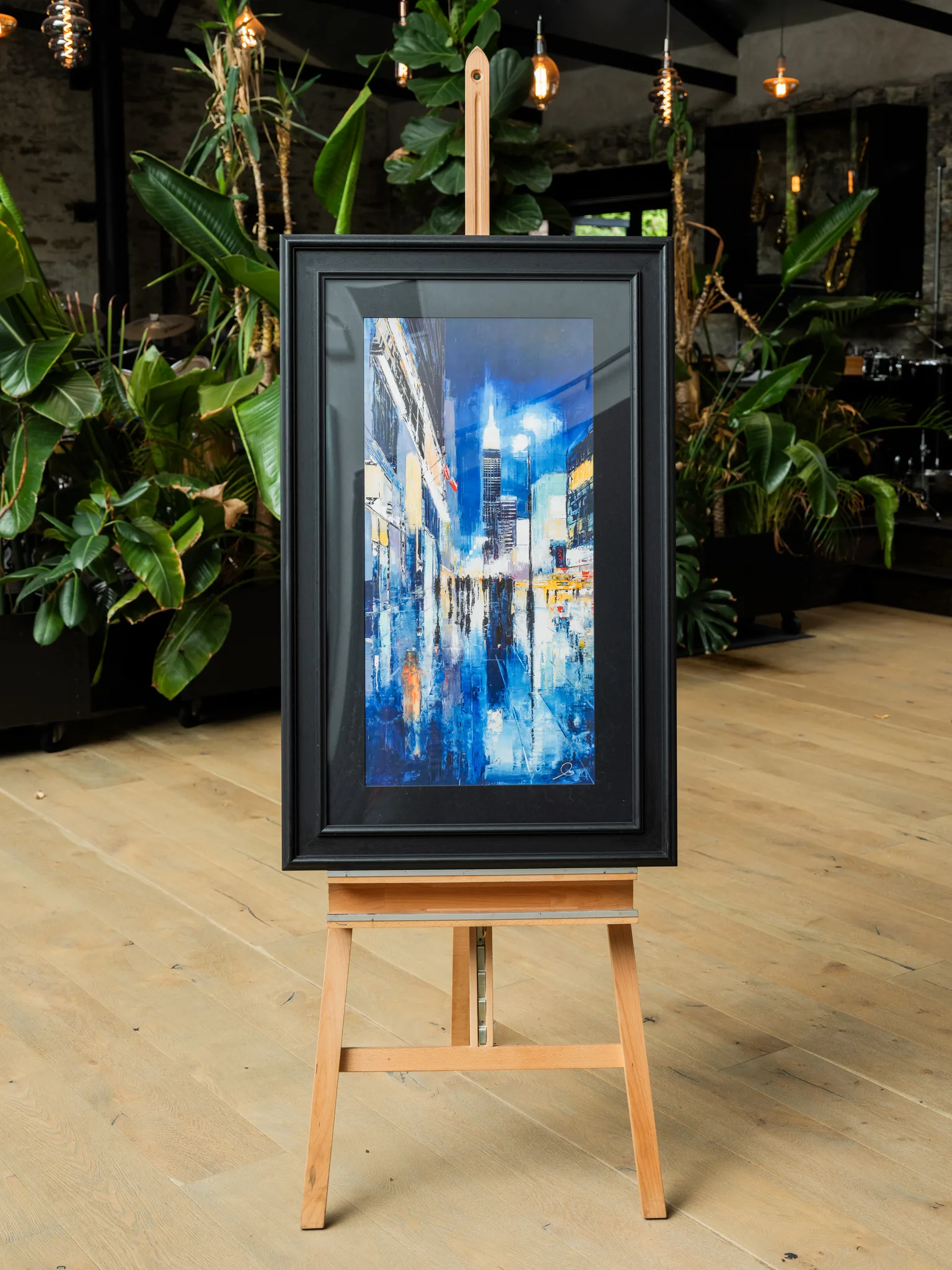 Electric City, a limited edition giclée print of New York at night, shown in a black frame on a wooden easel. A tall, illuminated skyscraper rises through glowing blue reflections on wet street tiles, with warm lights along the sides.