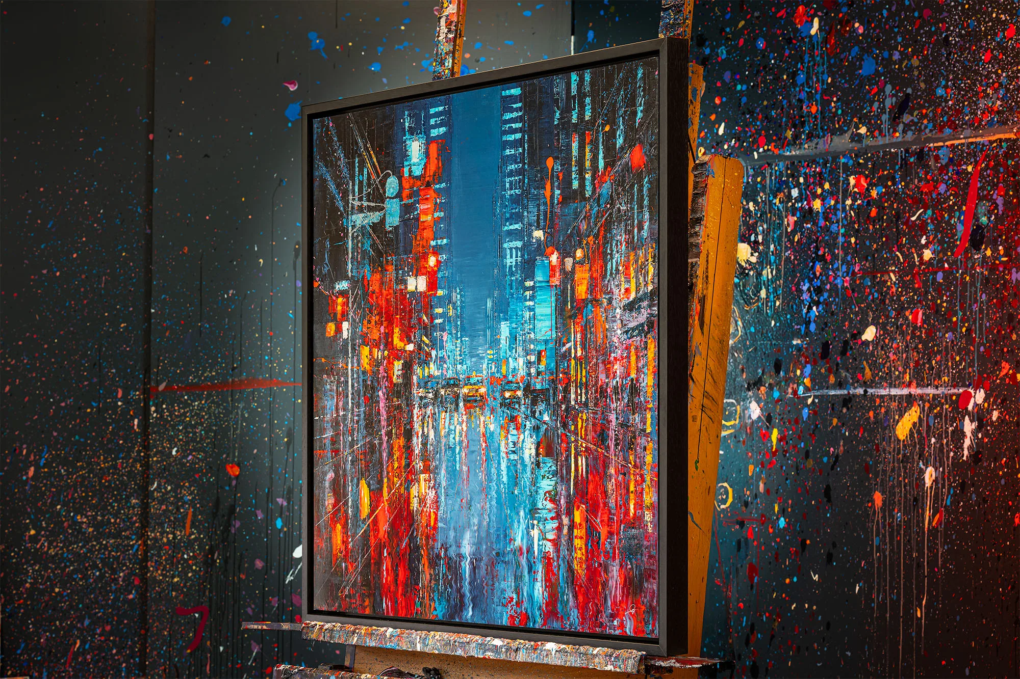 Electric Crossroads, a limited edition embellished giclée print on aluminium by Paul Kenton, shown in a dark frame in a paint-splattered studio. A night street of New York glows in red, orange and cyan, with towering lit buildings.