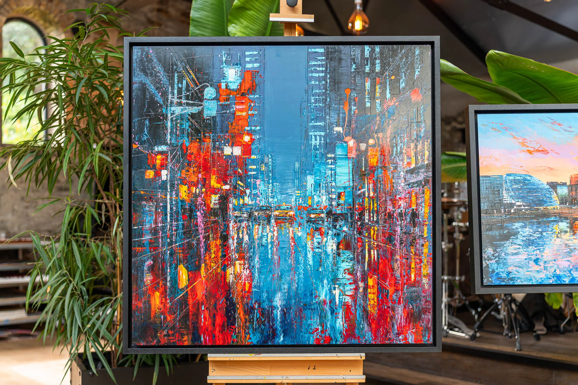 Electric Crossroads, a limited edition embellished giclée print by Paul Kenton, in a black frame on an easel in a plant-filled room. A bold New York street scene of blue, red and orange city lights reflected in wet pavement.