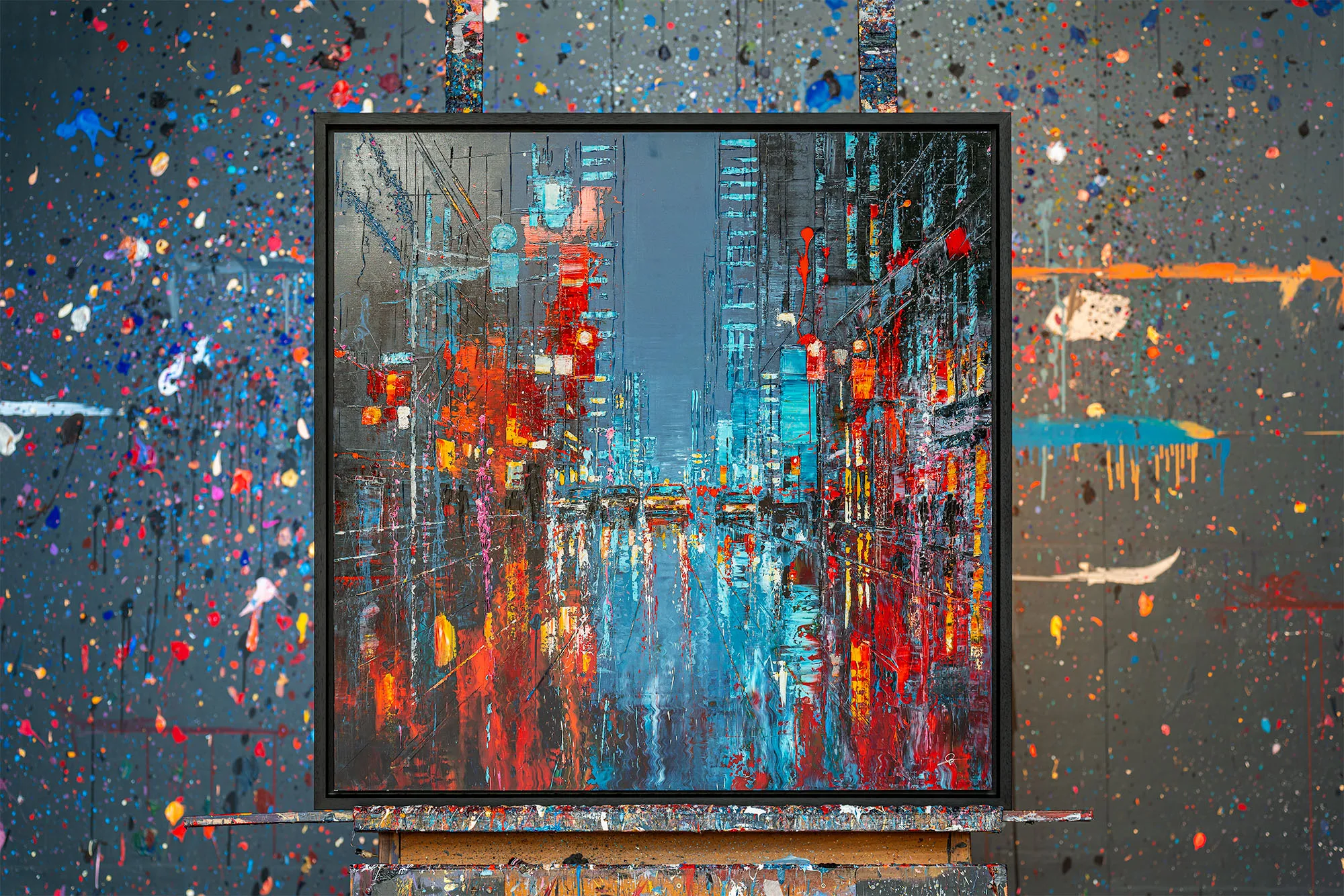 Electric Crossroads, a limited edition print by Paul Kenton, hangs on an easel in a paint-splattered studio, showing a rain-slicked New York street of blue towers and red and orange lights.