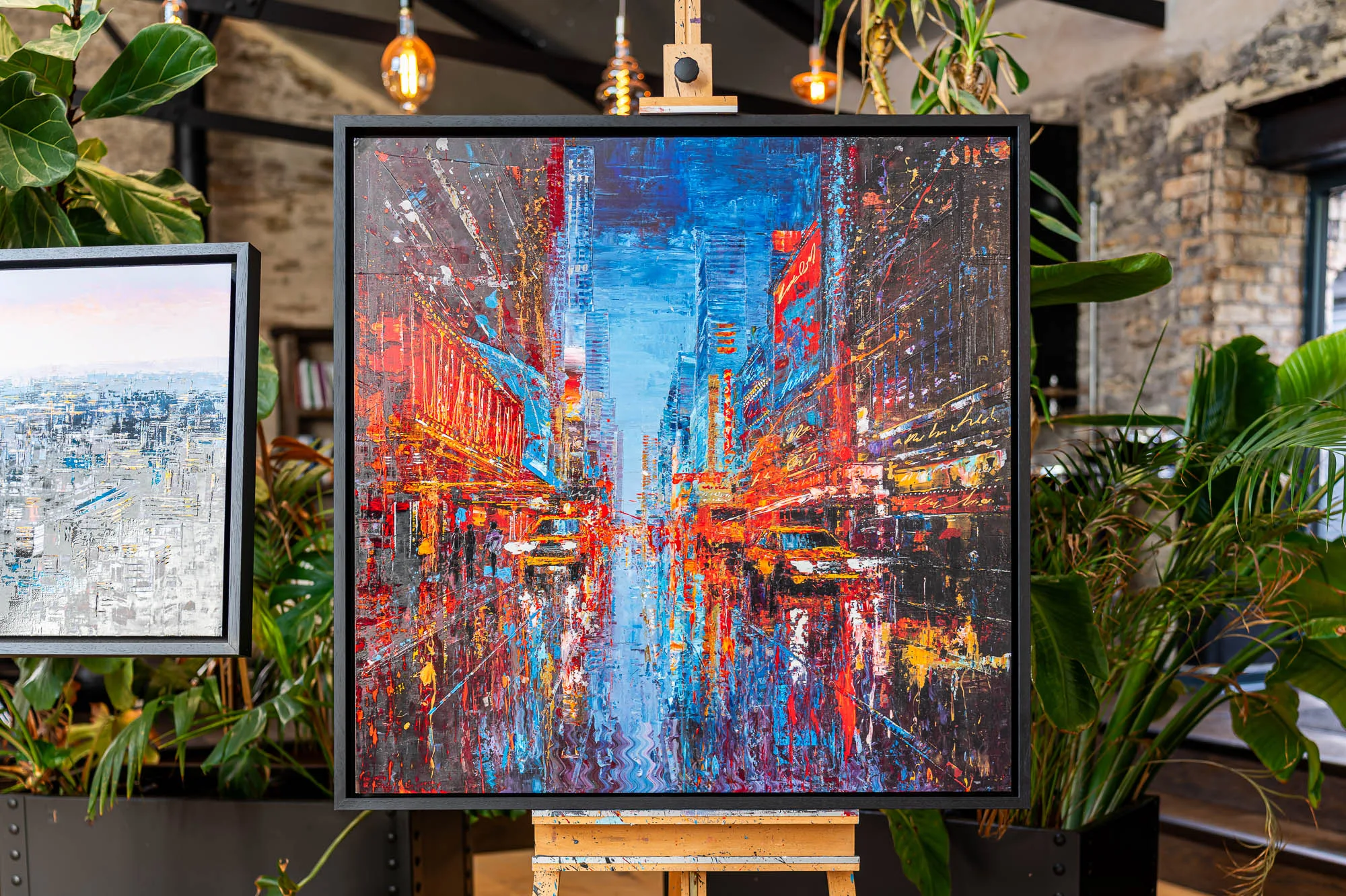 Empire City Spectrum, a limited edition print by Paul Kenton, hangs on an easel in a gallery, a night street scene of New York in red and blue neon with a dark frame.