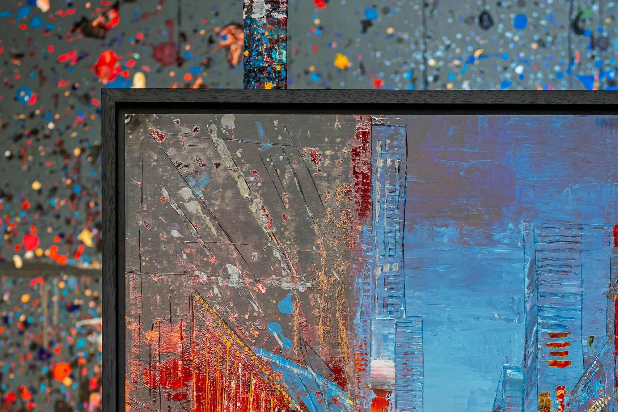 Close-up of Empire City Spectrum, a limited edition print in a black frame, showing thick textured paint in grey, red and blue. Raised blocky skyscraper forms rise against a blue sky, with a splattered backdrop behind.