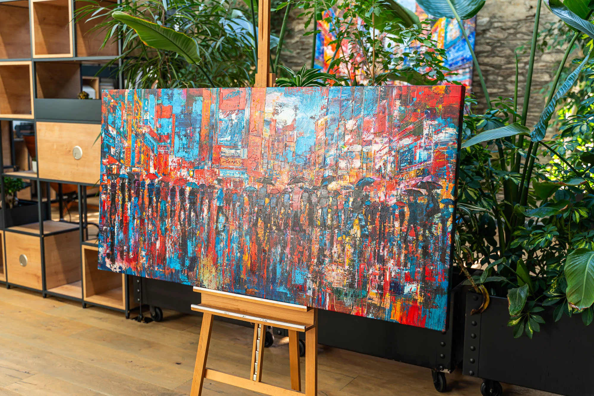 Fairytale Of New York, a limited edition canvas print, rests on a wooden easel among green plants, its red and blue street crowded with umbrellas.