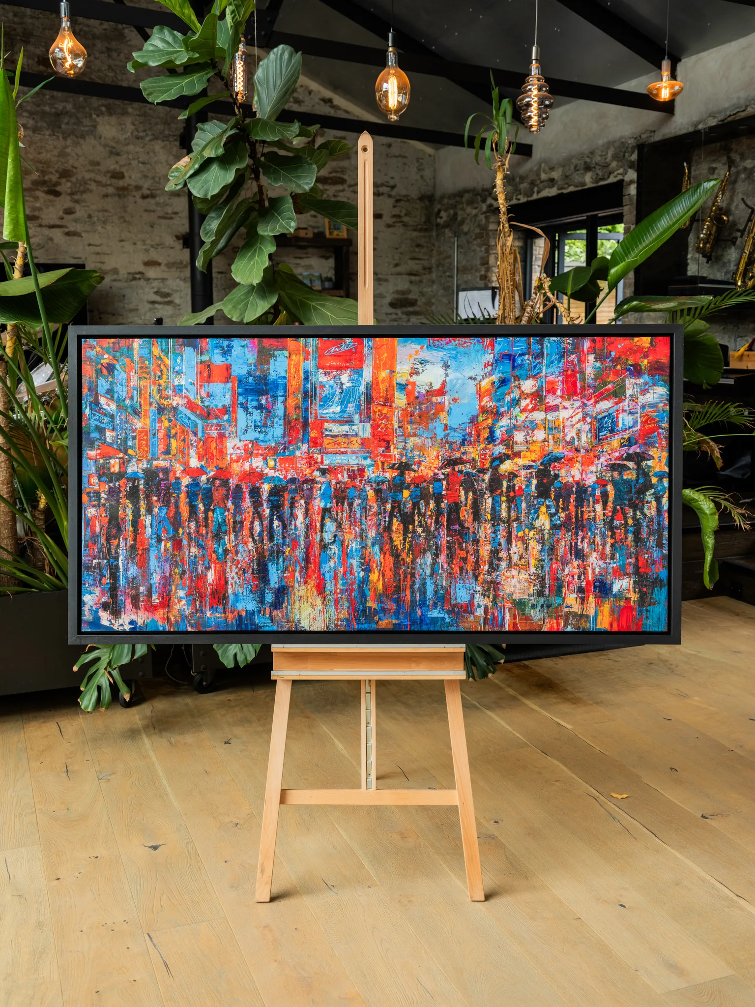 Fairytale Of New York, a limited edition print by Paul Kenton, on an easel in a rustic room. Bold red, orange and blue city street with blurred umbrella-carrying crowds and glowing signs.