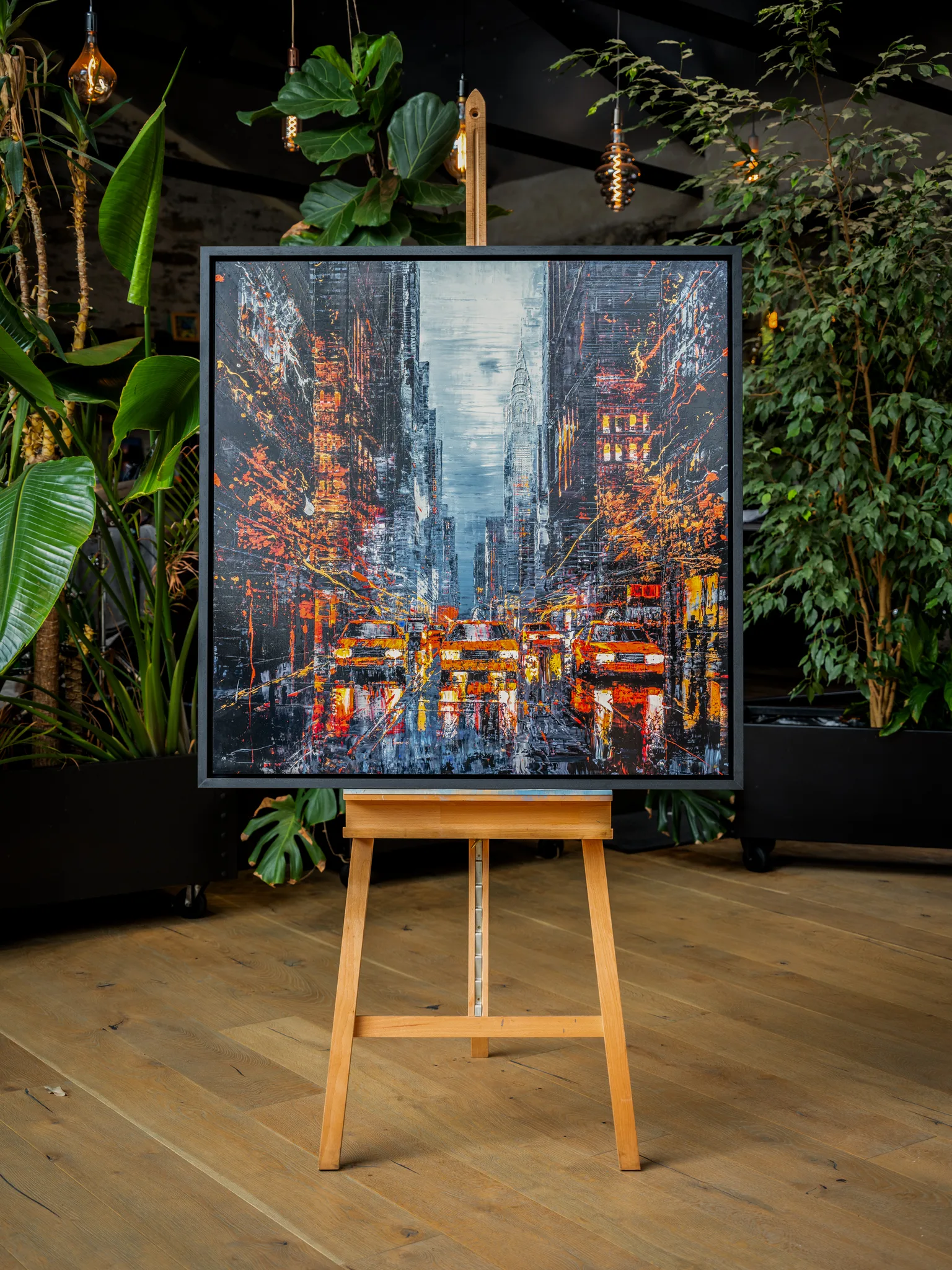 Flight of the Taxis, a limited edition print by Paul Kenton, displayed in a black frame on a wooden easel among potted plants and hanging Edison bulbs. A bold, textured New York street scene shows yellow taxis glowing in red and amber light.