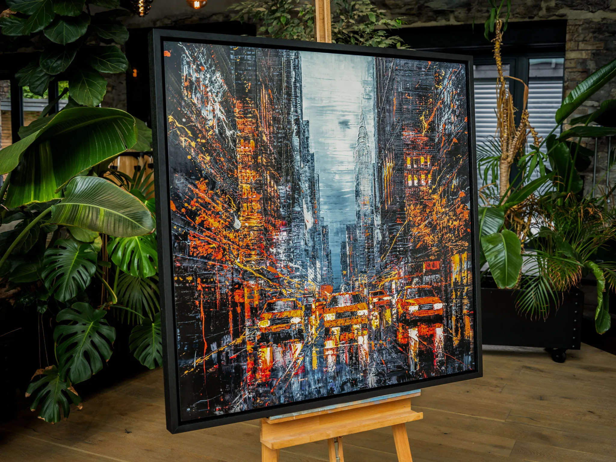 Flight of the Taxis, a limited edition print by Paul Kenton, in a black frame on a wooden easel among large green plants. A bold, textured night street scene of New York with glowing orange taxis and tall dark towers.