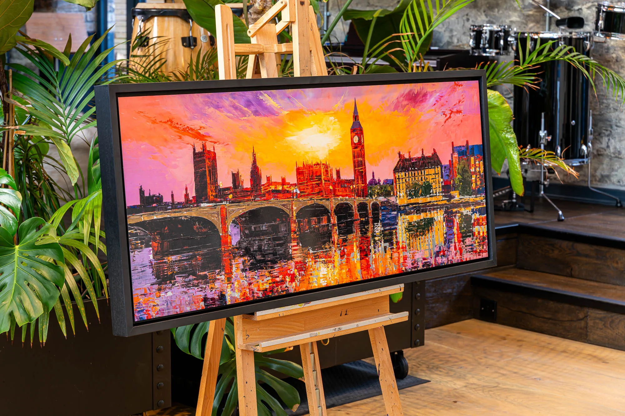 London Ablaze, a limited edition print by Paul Kenton, hangs in a black frame on a wooden easel in a plant-filled room. It shows the Houses of Parliament and Big Ben at sunset, in fiery oranges and pinks.