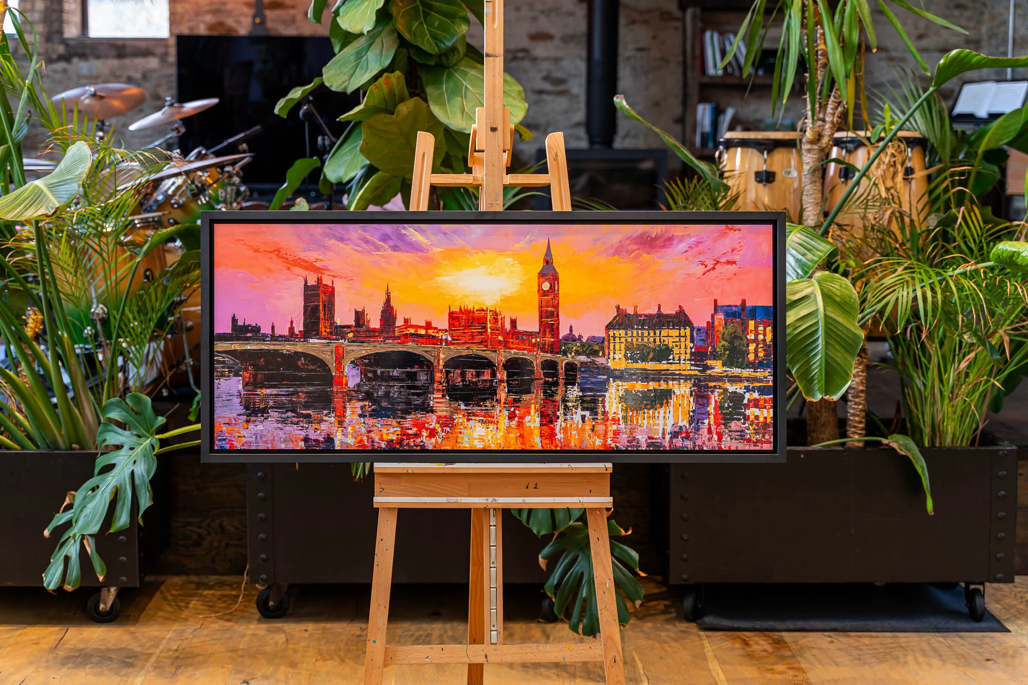 London Ablaze, a limited edition print by Paul Kenton, in a black frame on a wooden easel in a plant-filled studio. The sunset cityscape shows Big Ben and a bridge over the Thames in magenta, orange and gold.