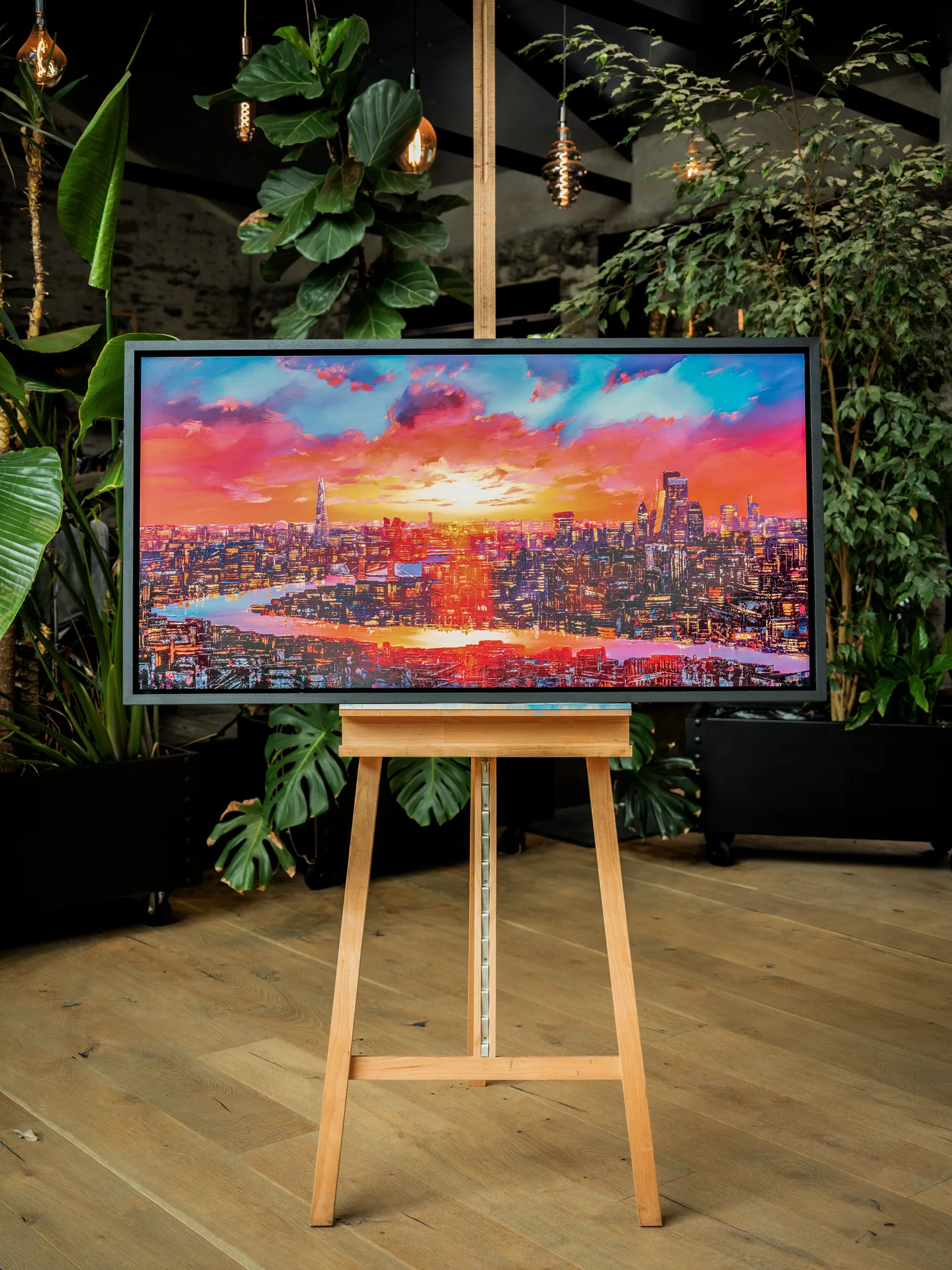 London Embers, a limited edition print by Paul Kenton, hangs in a frame on a wooden easel amid green plants, showing a city skyline at sunset in fiery reds, oranges and blues.