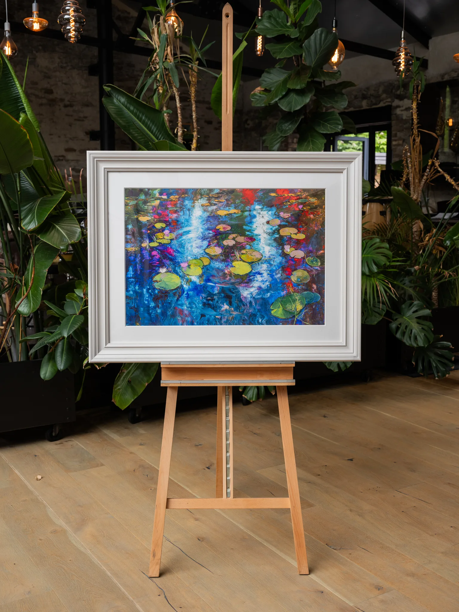 Lily Light, a limited edition print by Paul Kenton, shown in a cream frame on a wooden easel in a plant-filled room, with blue water, pads and red and pink flecks.
