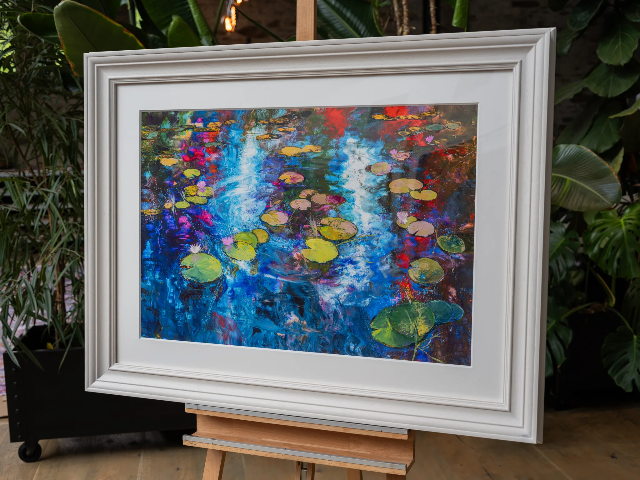 Lily Light, a limited edition print by Paul Kenton, in a white frame on an easel: bold blue, red and green water with floating lily pads and pale blossoms, set among indoor plants.