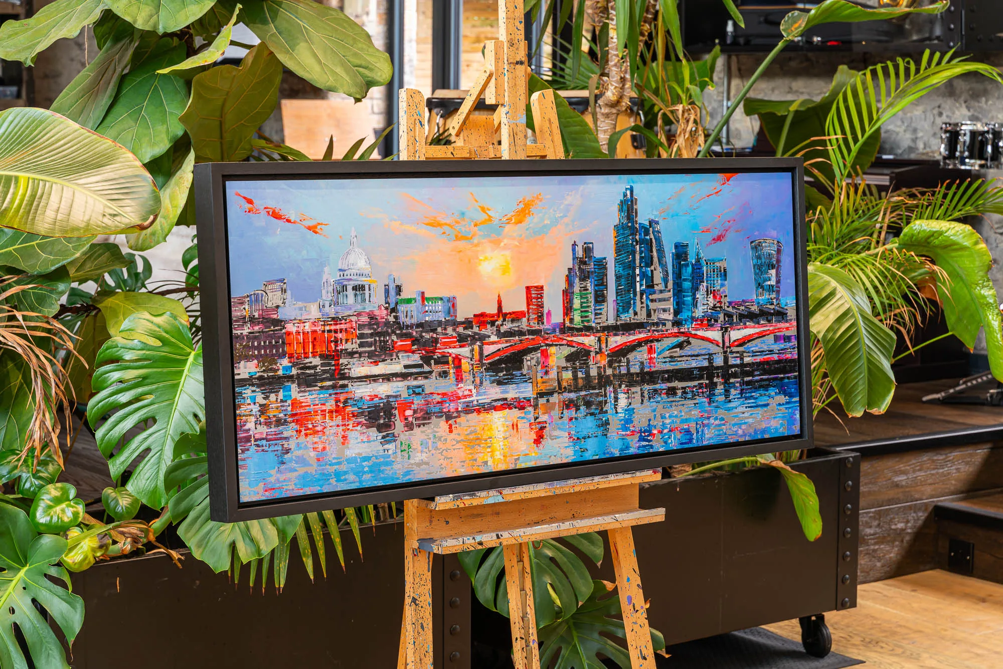 London Reflects, a limited edition print by Paul Kenton, shown in a black frame on a wooden easel among large green plants. It shows a dusky London skyline with a domed cathedral, glass towers and a red arched bridge over water, in bold blue, orange and red.
