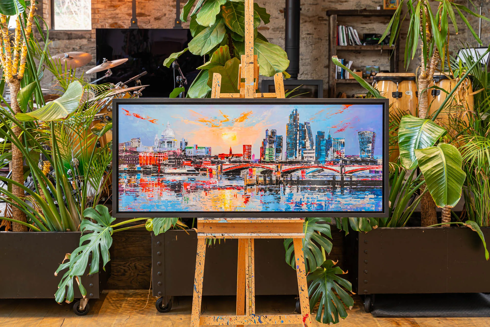 London Reflects, a limited edition print by Paul Kenton, hangs in a black frame on an easel amid lush plants. It shows St Paul's dome and city towers at sunset, with fiery orange and blue reflections on the Thames.