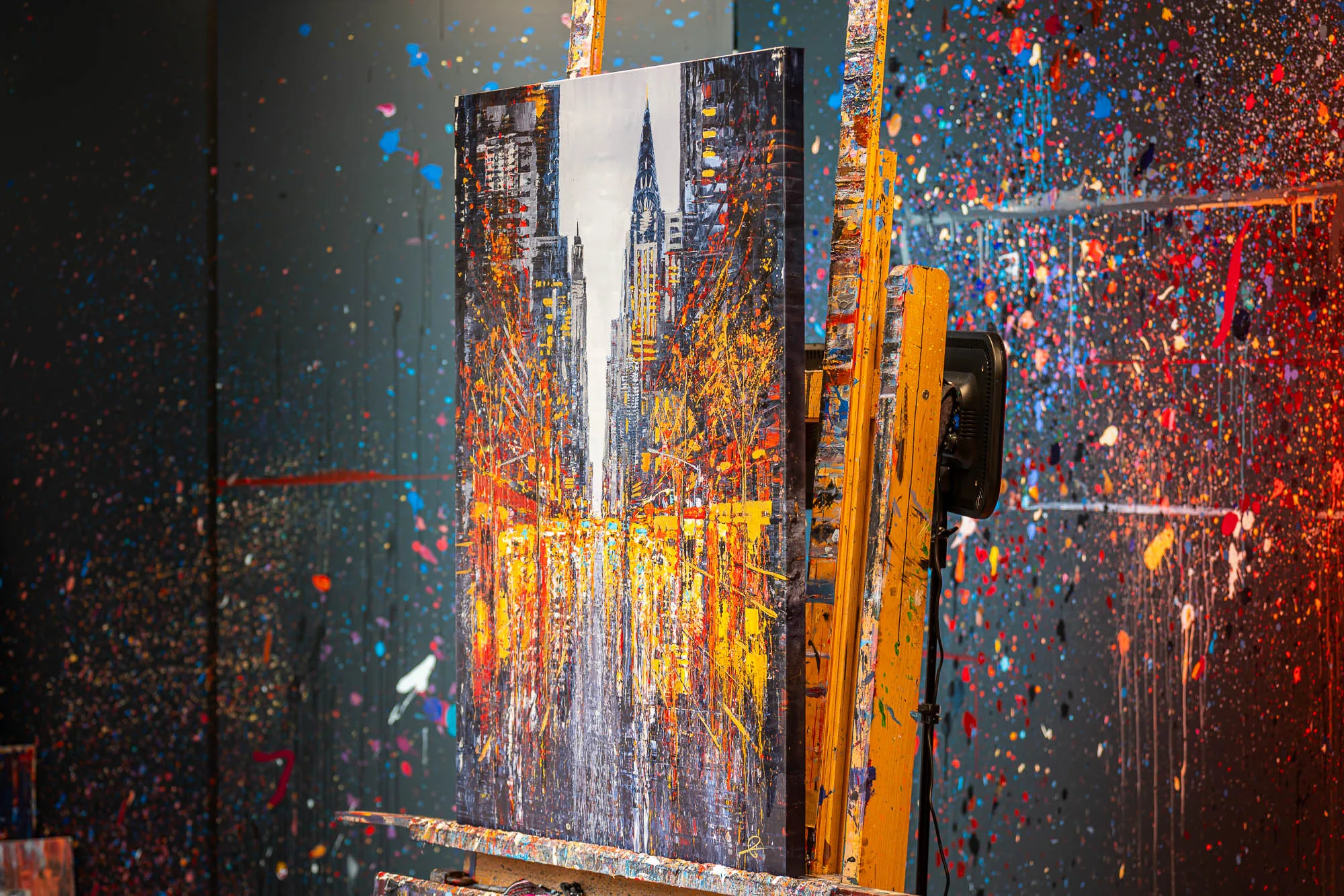 New York Serenade, an embellished giclée print on boxed canvas, on a yellow easel in a studio splattered with paint, showing a tall city tower lit in gold and orange against dark buildings.