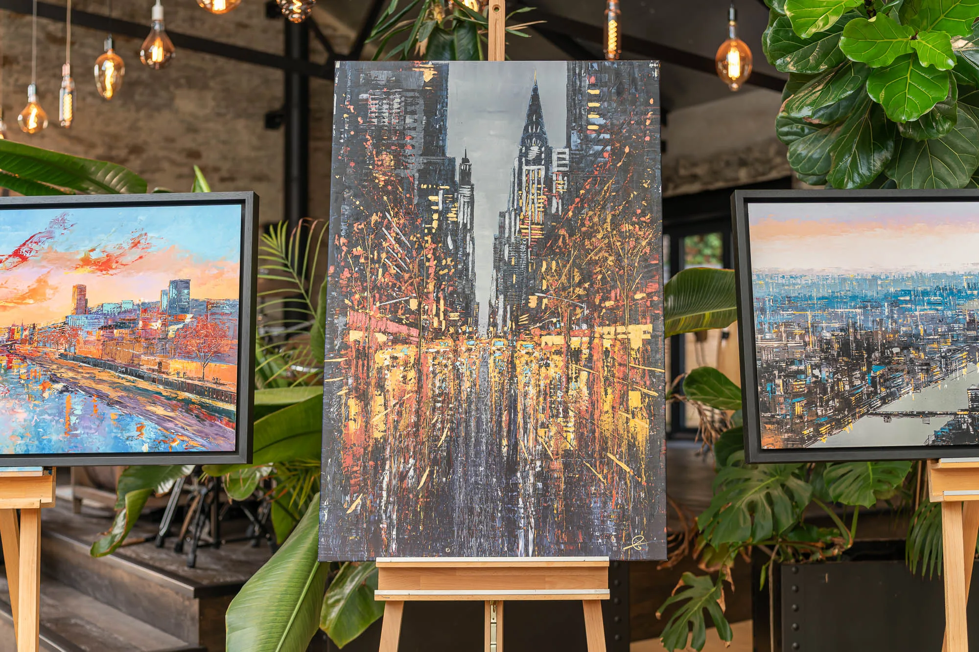 New York Serenade, a limited edition embellished giclée print on canvas, hangs on a wooden easel in a room with trailing plants and bulb lights, flanked by two framed cityscapes. Its dark street glows with amber and gold streaks beneath towering skyscrapers.
