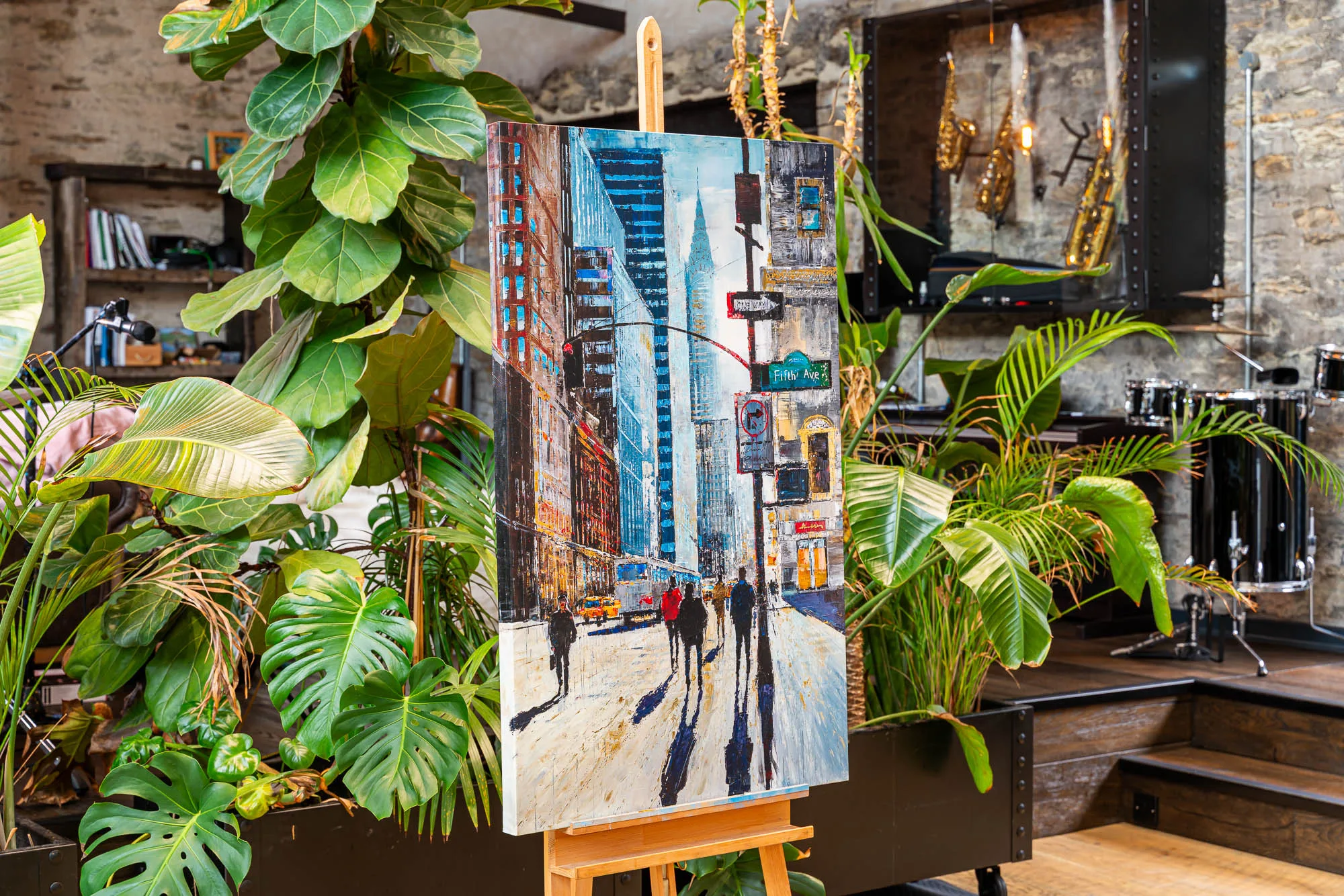 New York Shadows, an embellished giclée print on canvas, on an easel among tall plants in a stone-walled room; a Fifth Avenue street scene of figures casting long shadows, in cool blues and warm ochres.