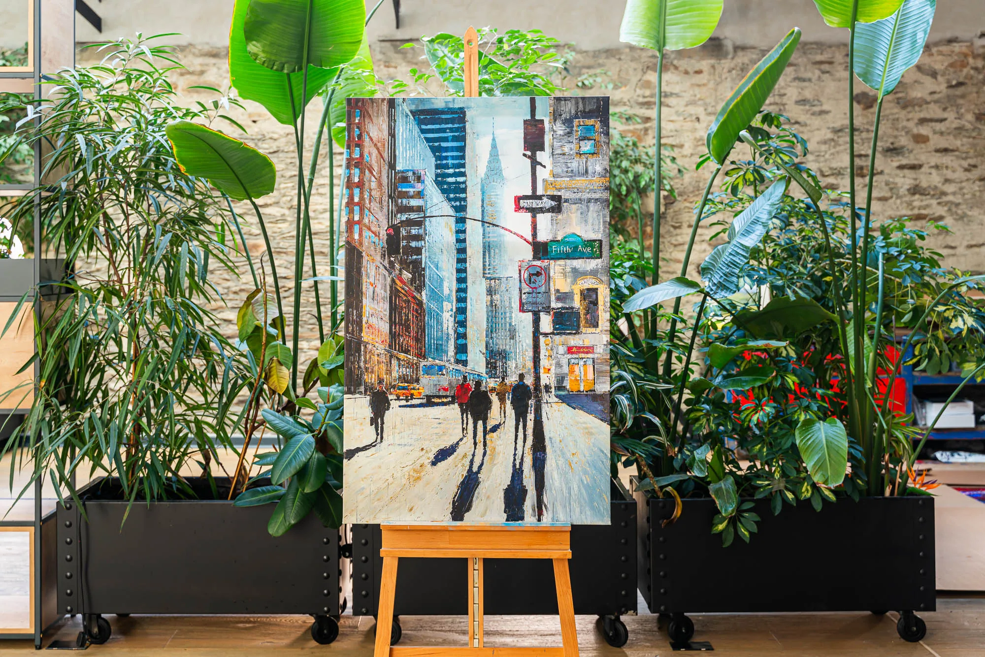 New York Shadows, an embellished giclée print by Paul Kenton on an easel among tropical plants: a Manhattan street of tall buildings, pedestrians casting long shadows, in cool blues and warm amber.