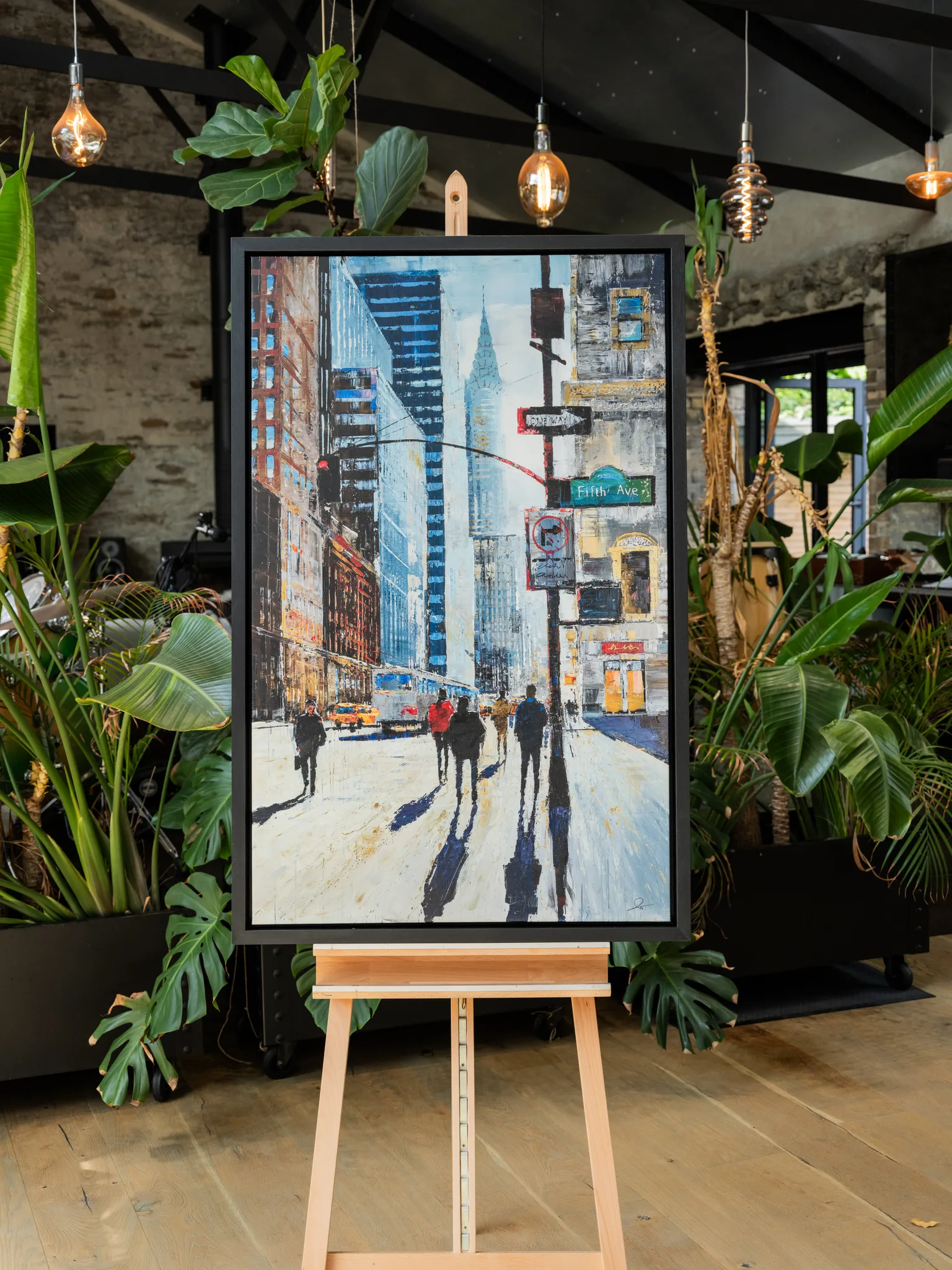 New York Shadows, a limited edition print by Paul Kenton on a black-framed canvas, displayed on a wooden easel in a plant-filled room. It shows a bustling Fifth Avenue street in blue and cream tones, with long shadows of pedestrians.