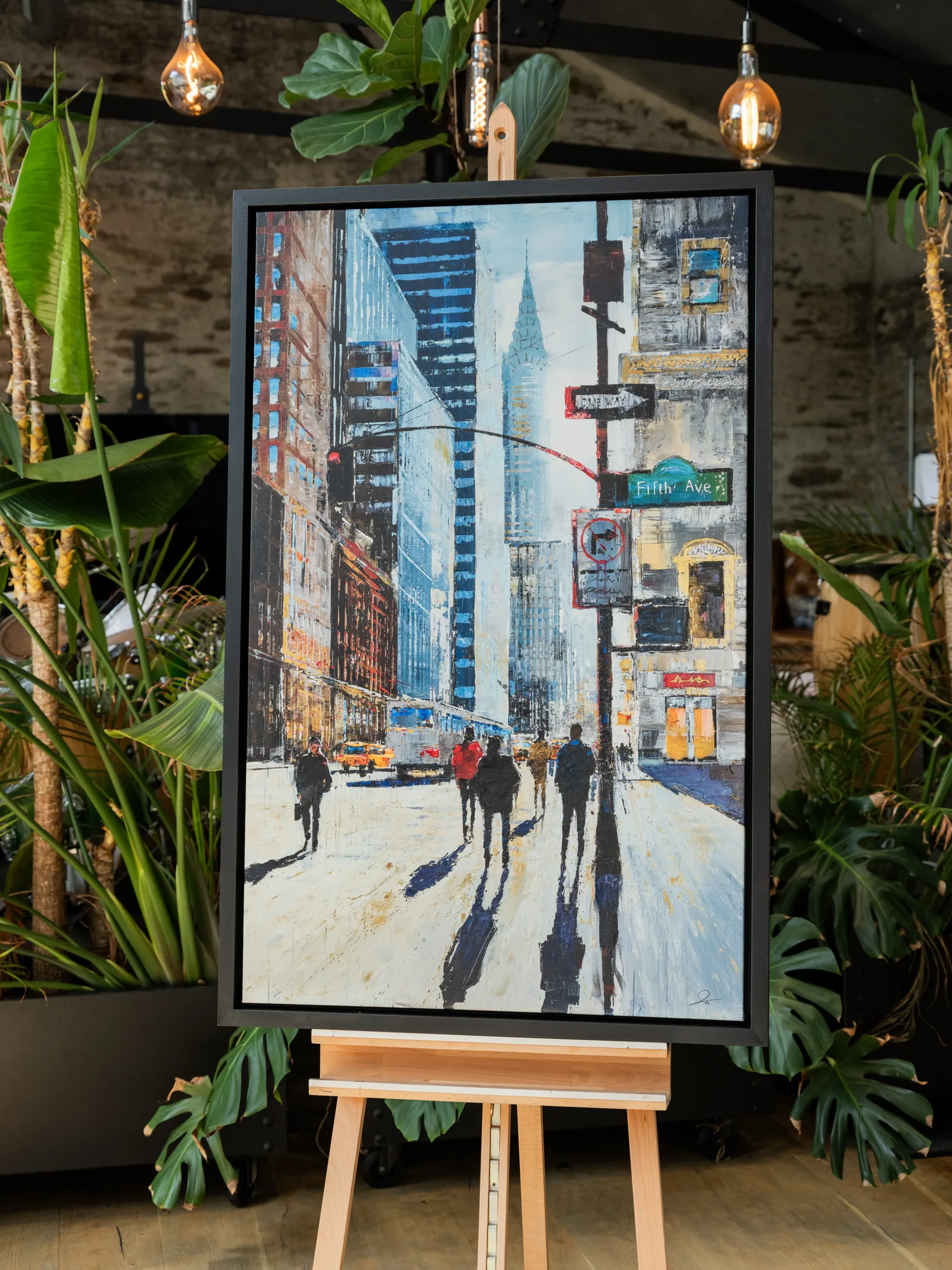New York Shadows, a limited edition canvas print by Paul Kenton, in a black frame on a wooden easel among green plants: a bustling Fifth Avenue street scene of walkers casting long shadows, with blue-grey towers and a yellow taxi.