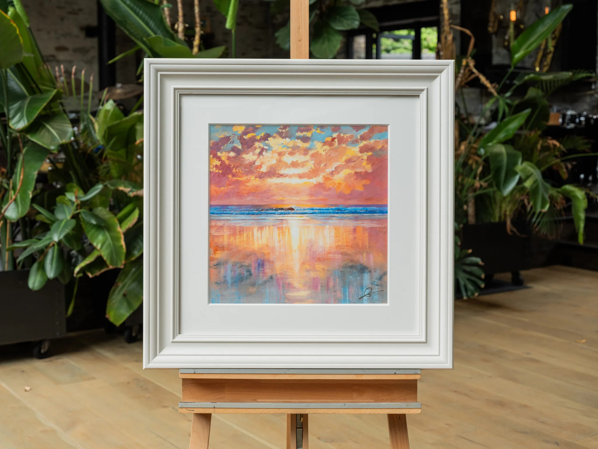 Ocean Harmony, a limited edition print by Paul Kenton, in a cream frame on a wooden easel in a room with leafy plants. A bold sunset over calm sea, with orange and pink clouds and warm light reflected on the water.