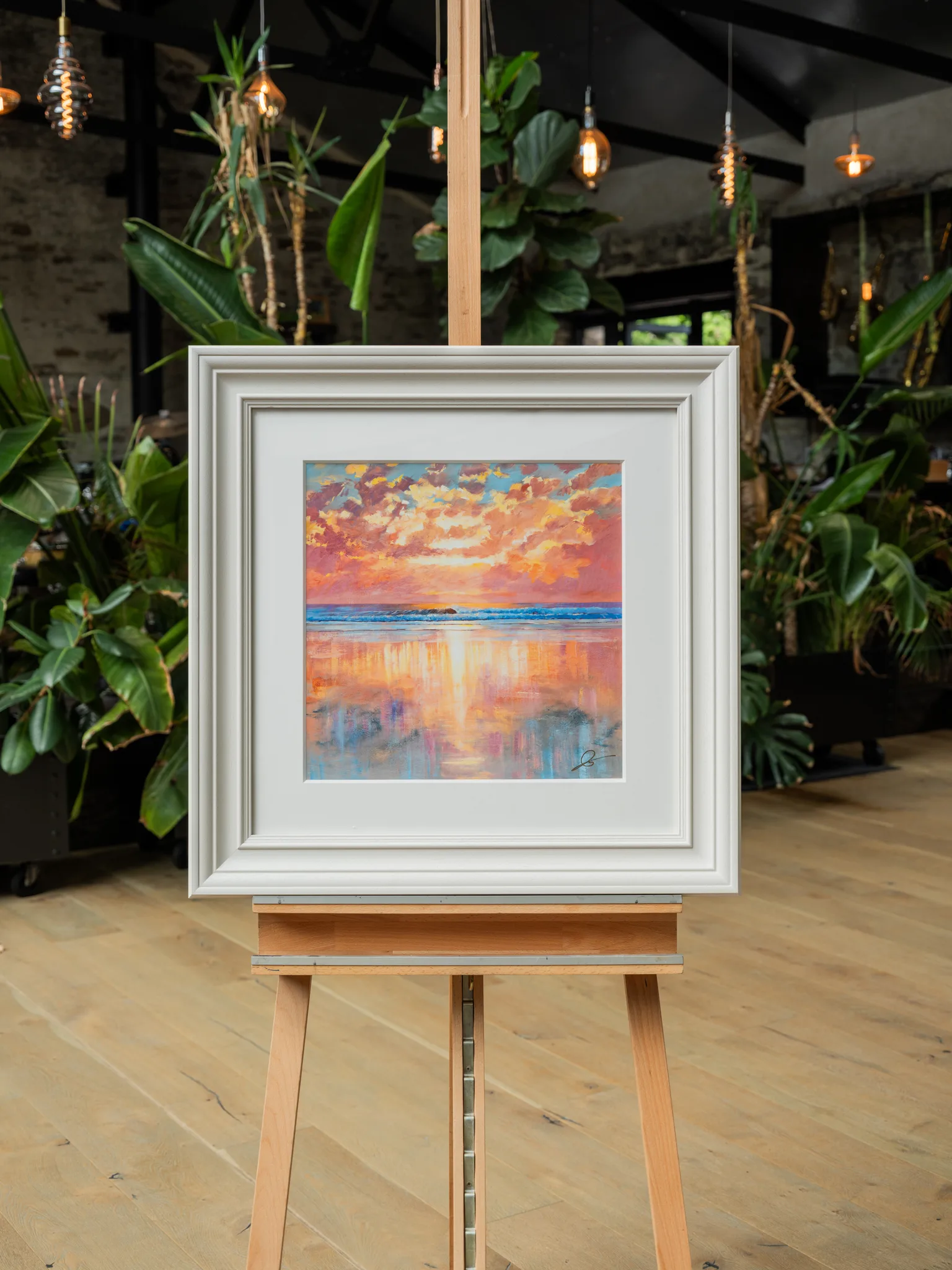 Ocean Harmony, a limited edition print by Paul Kenton, hangs in a cream frame on a wooden easel in a plant-filled room, showing a glowing orange and pink sunset over calm water.