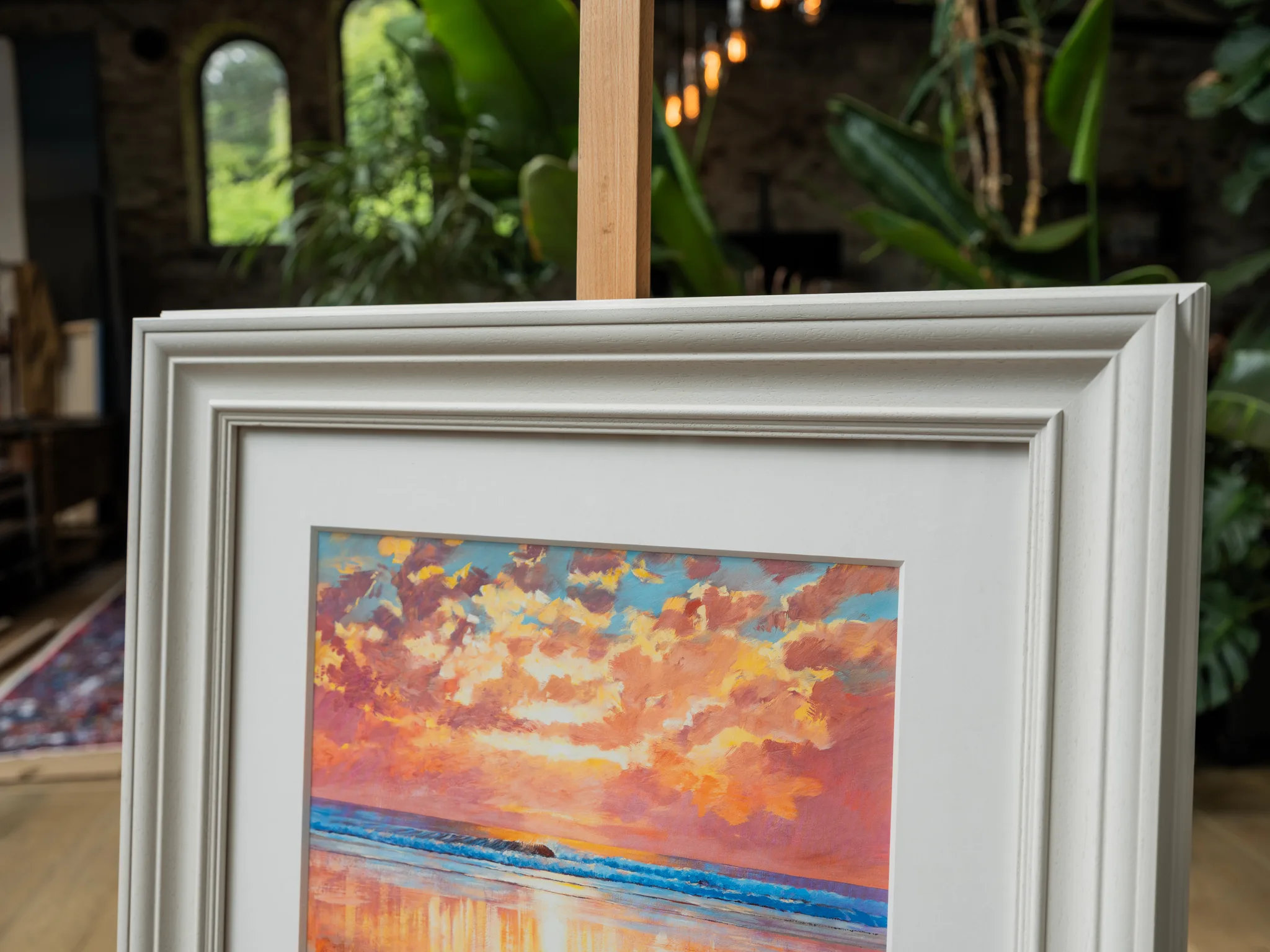 Ocean Harmony, a limited edition giclée print by Paul Kenton, in a white frame on an easel in a room with plants and an arched window. It shows a bold sunset sky of pink, orange and gold clouds above a blue sea.