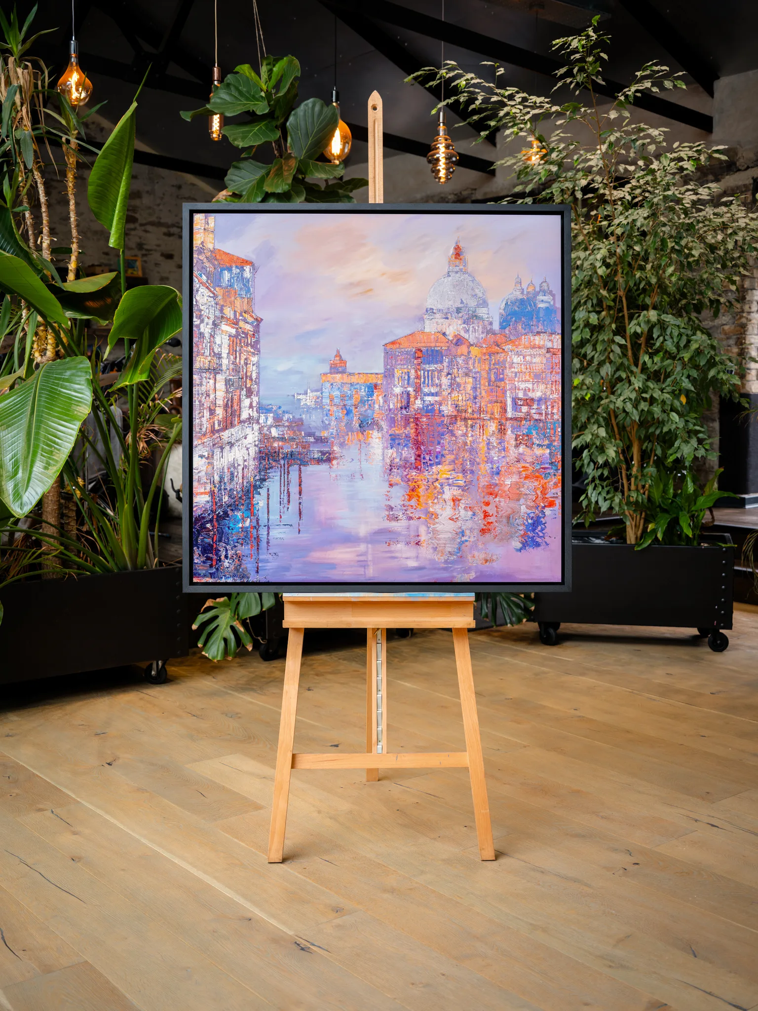 Opalescent Venice, a limited edition print by Paul Kenton, hangs in a plant-filled room on a wooden easel, showing a textured canal scene with a domed church in lilac, peach and orange.