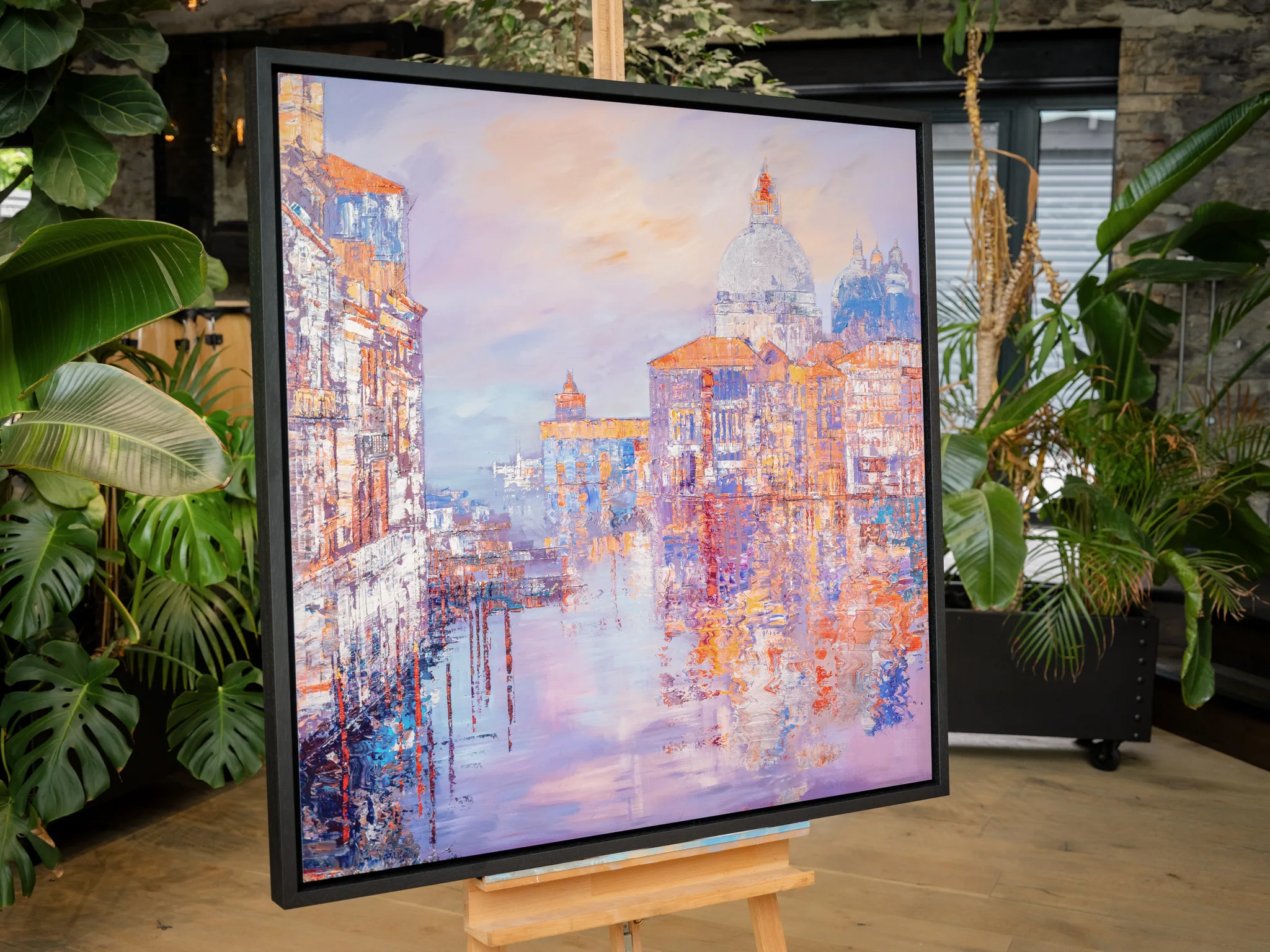 Opalescent Venice, a limited edition print in a black frame on a wooden easel amid lush plants, its lilac and peach canal scene glowing.