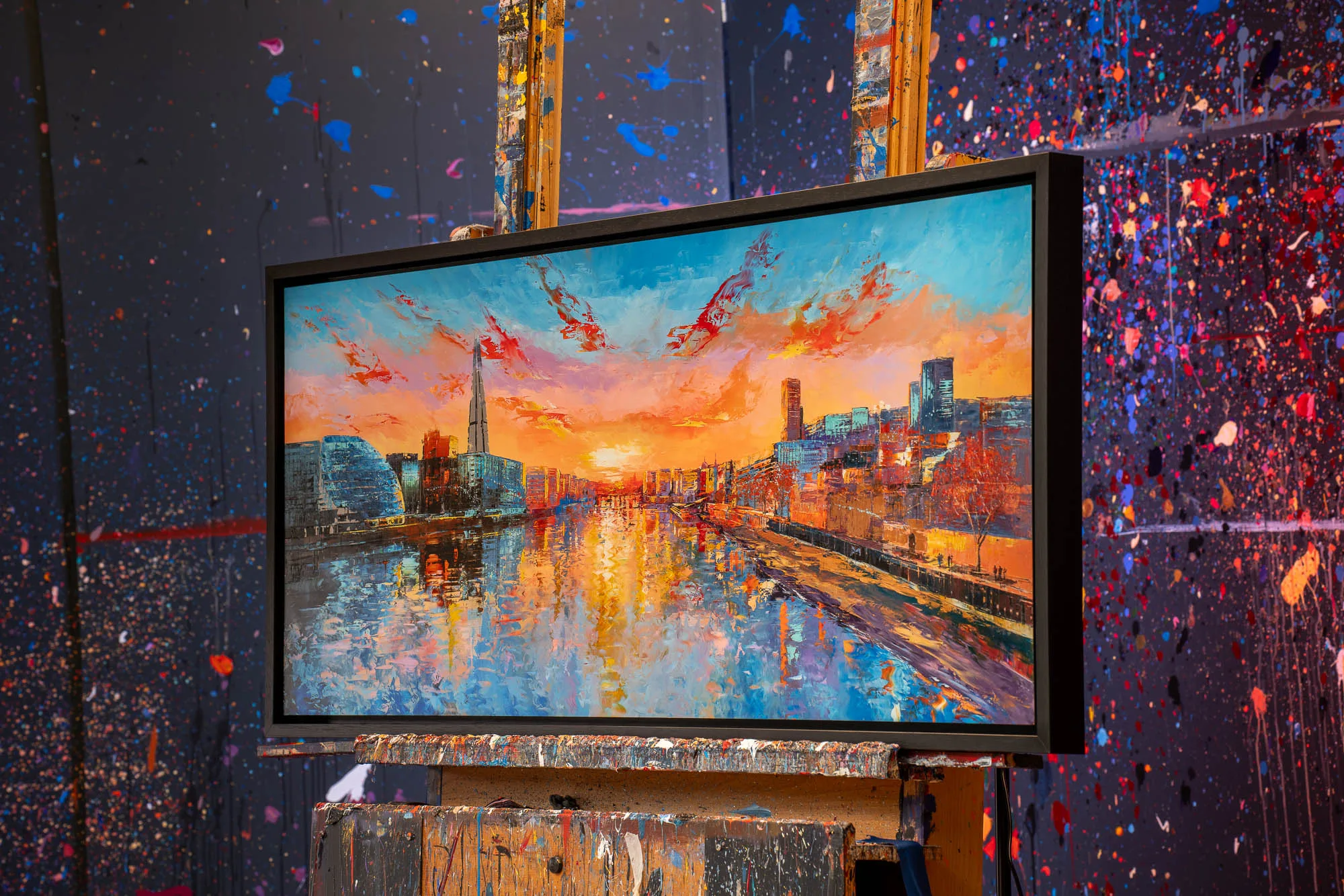 Panoramic Glow, a limited edition print by Paul Kenton, sits on a paint-splattered studio easel, showing a London river at sunset under orange skies.