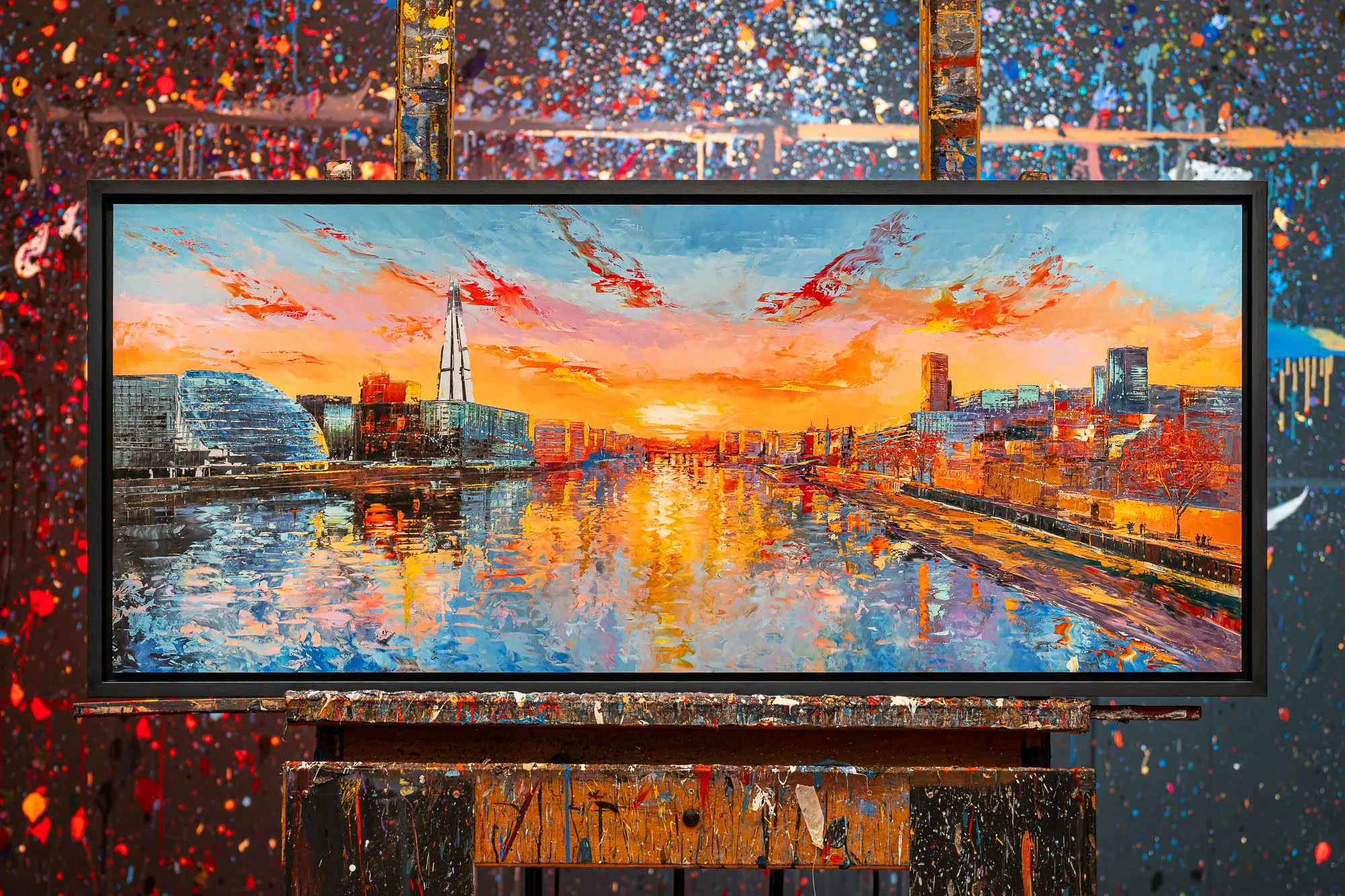 Panoramic Glow, a limited edition print by Paul Kenton, hangs in a black frame on an easel: a London riverside at sunset with orange skies, a glowing reflective river and a tall glass tower.