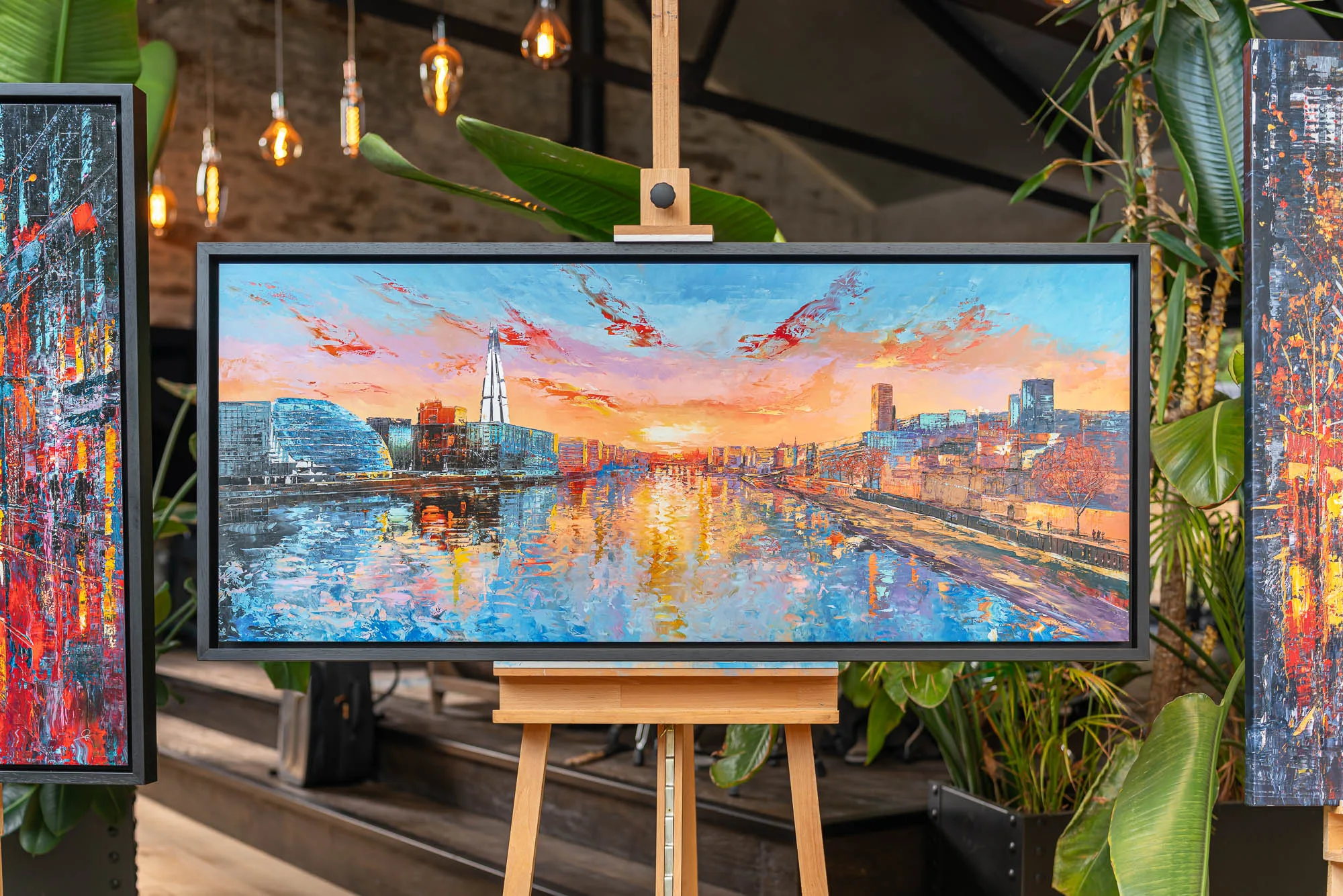 Panoramic Glow, a limited edition print by Paul Kenton, shown in a dark frame on a wooden easel in a warm café. It depicts London's river skyline at sunset, with a glowing orange and blue sky and golden reflections on the water.