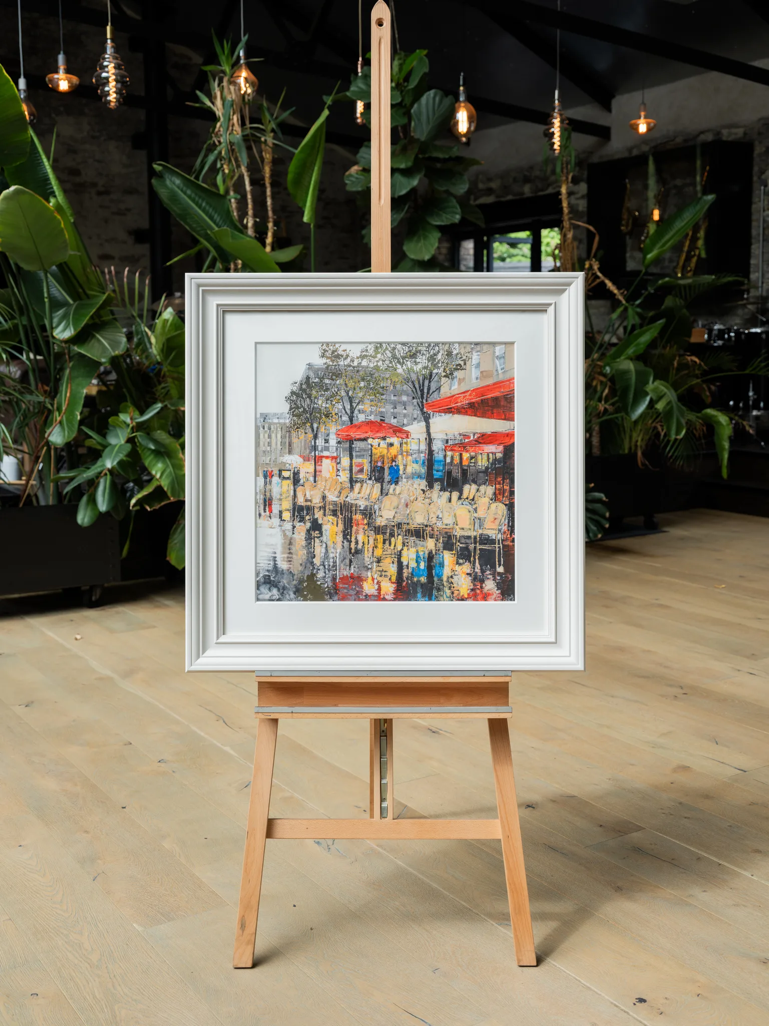 Parisien Life, a limited edition print by Paul Kenton, hangs in a white frame on a wooden easel in a plant-filled room. It shows a Paris café terrace with red awnings and rows of chairs.