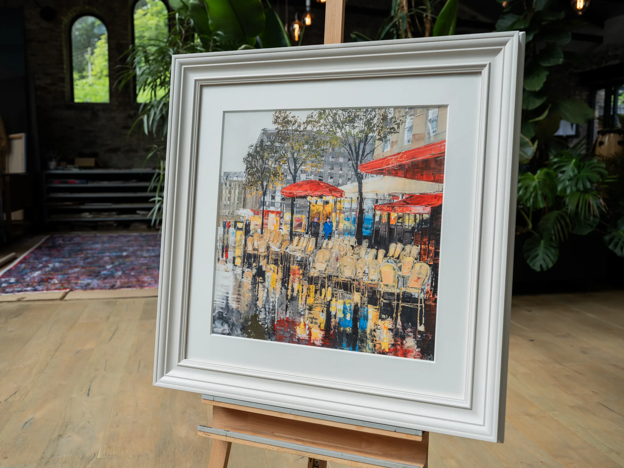 Parisien Life, a limited edition print by Paul Kenton, hangs in a white frame on an easel in a bright room. The textured street scene shows red-canopied café tables and chairs reflected in wet pavement.