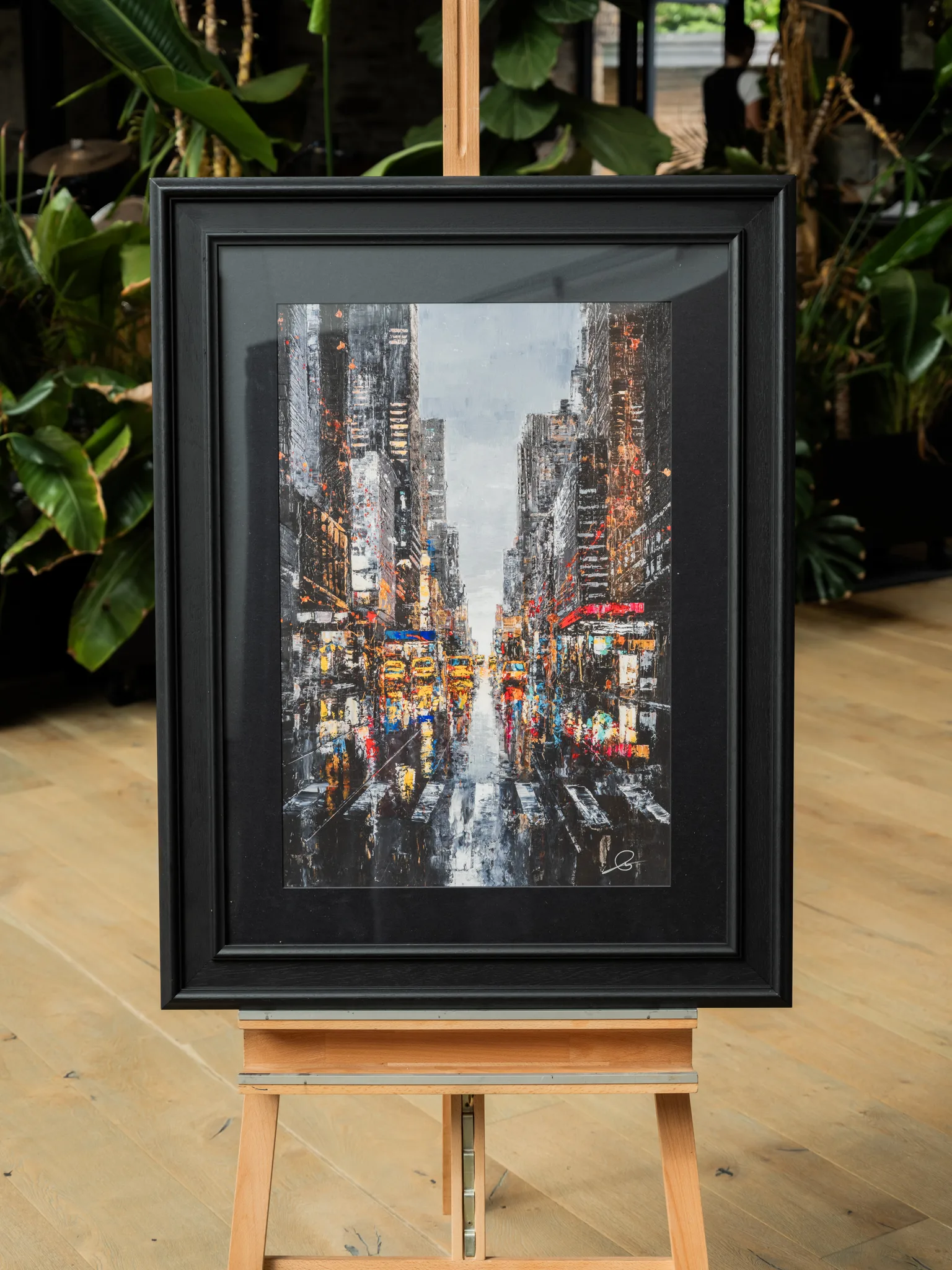 Shades of the City, a limited edition giclée print by Paul Kenton, hangs in a black frame on a wooden easel, showing a rain-slick New York street of glowing yellow and red lights.