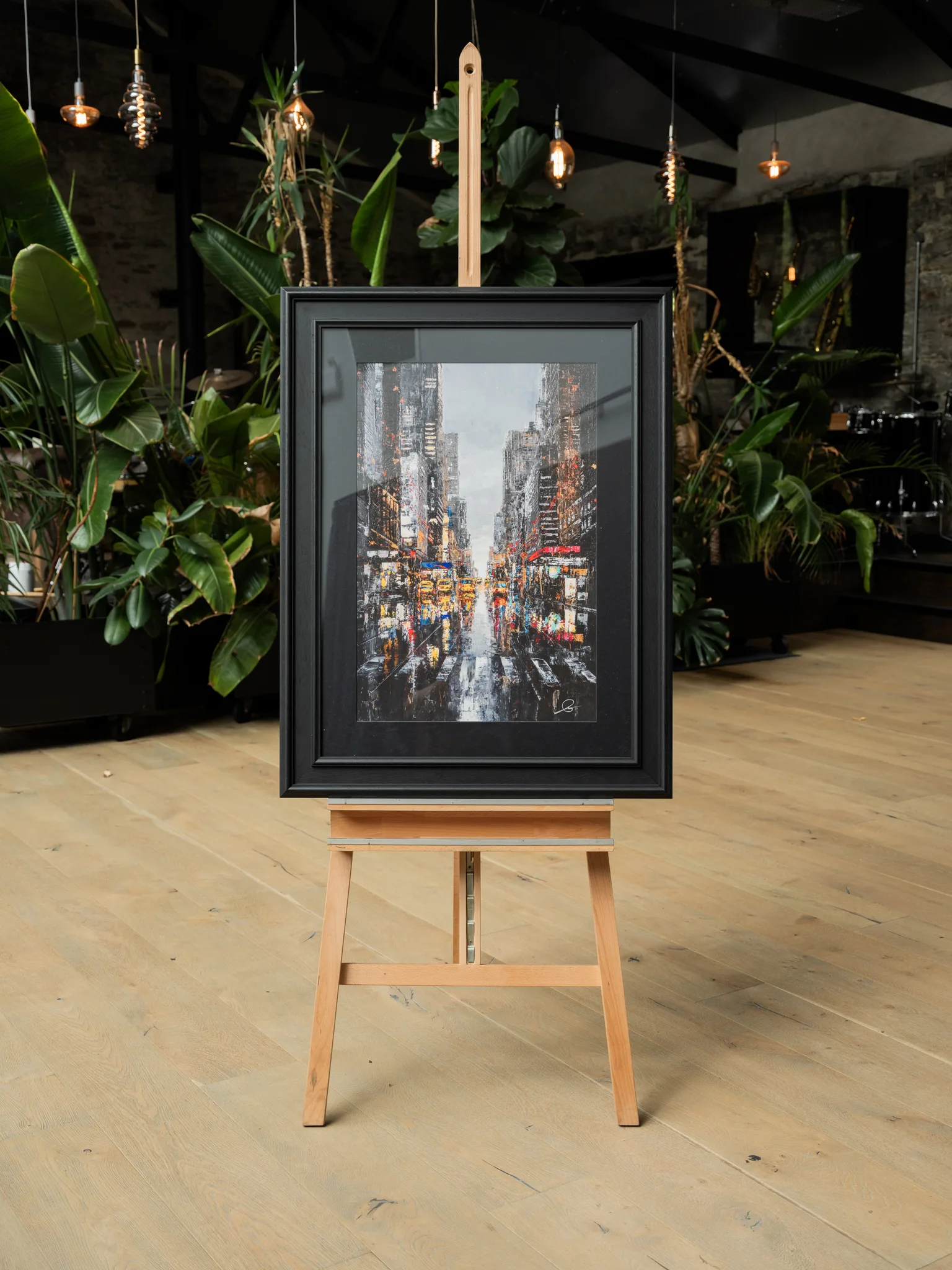 Shades of the City, a limited edition print of New York, framed in black on a wooden easel among tropical plants and hanging Edison bulbs. A rainy street scene with glowing yellow and red lights.