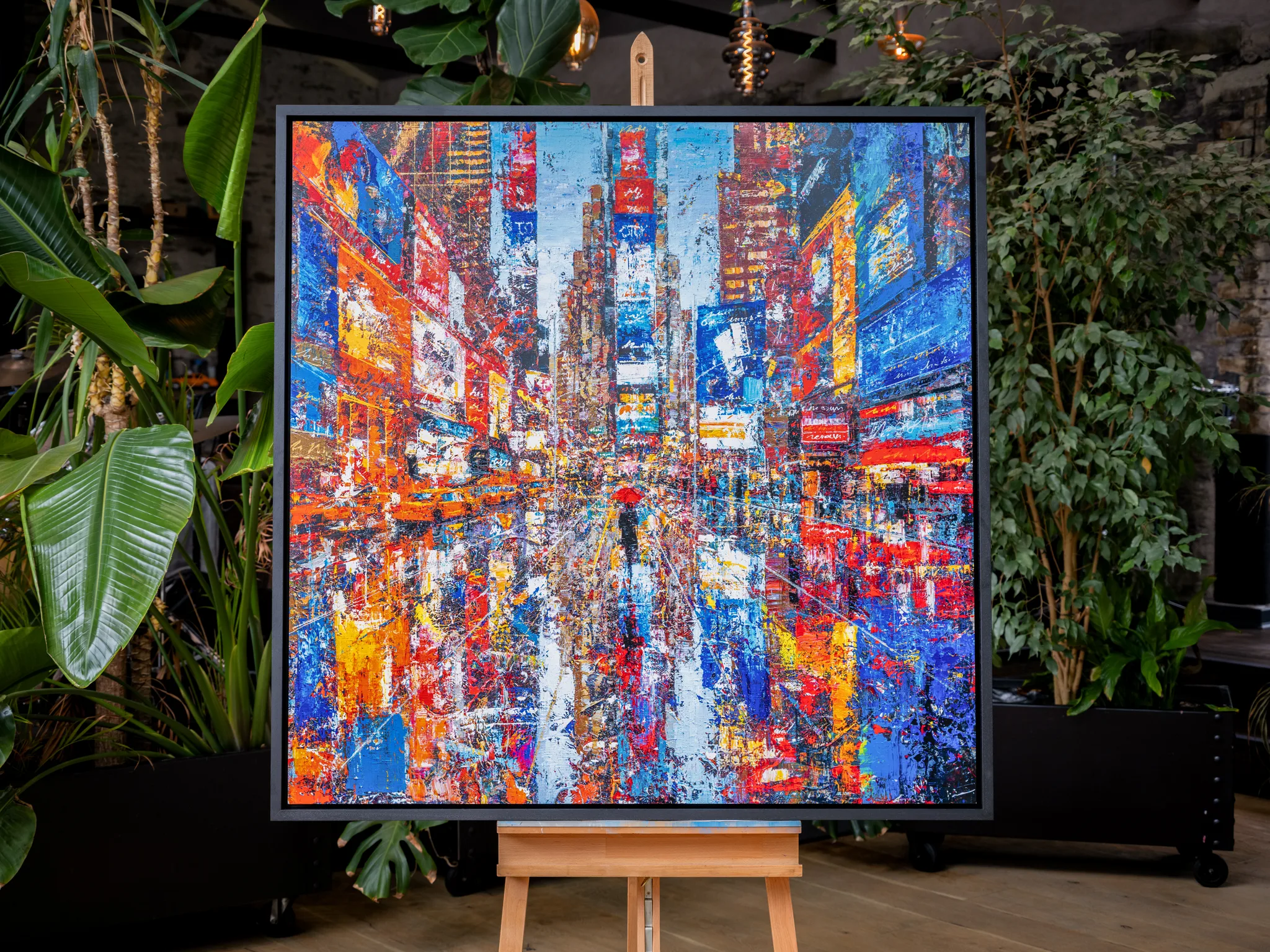 Steps Through Times Square, a limited edition print by Paul Kenton, hangs in a framed black border on a wooden easel among green indoor plants. Bold blue, red and orange city lights fill the textured street scene.