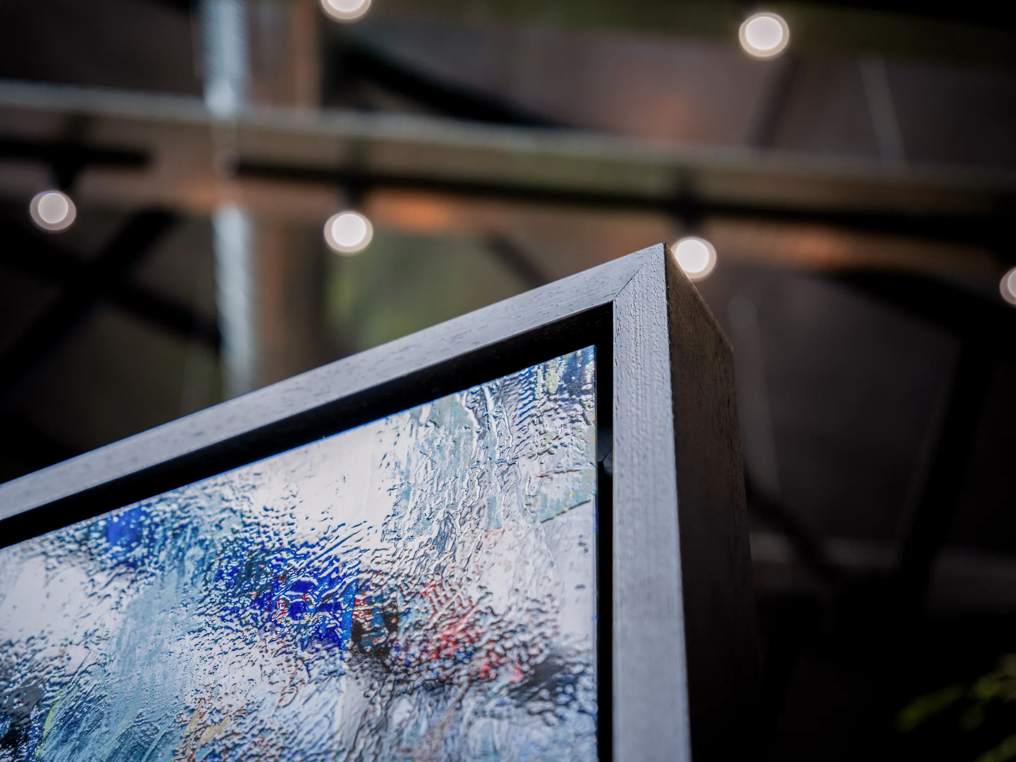 Close-up of the dark wooden frame corner of Steps Through Times Square, a limited edition print by Paul Kenton, showing textured blue, white and red brushwork beneath a glossy surface, with blurred string lights behind.