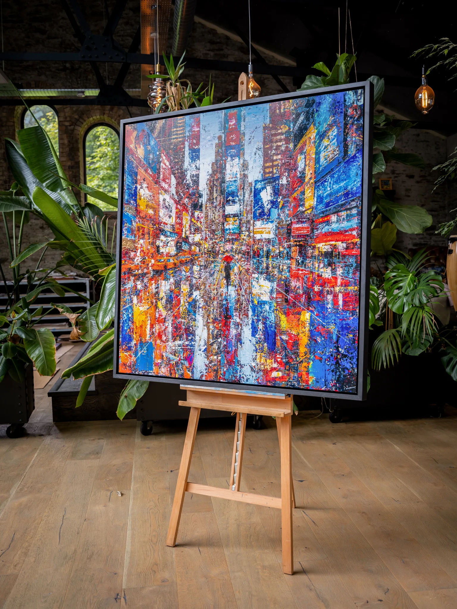 Steps Through Times Square, an embellished giclée print on aluminium by Paul Kenton, on a wooden easel in a plant-filled room, showing a bold, textured city street with glowing red, blue and orange signs.