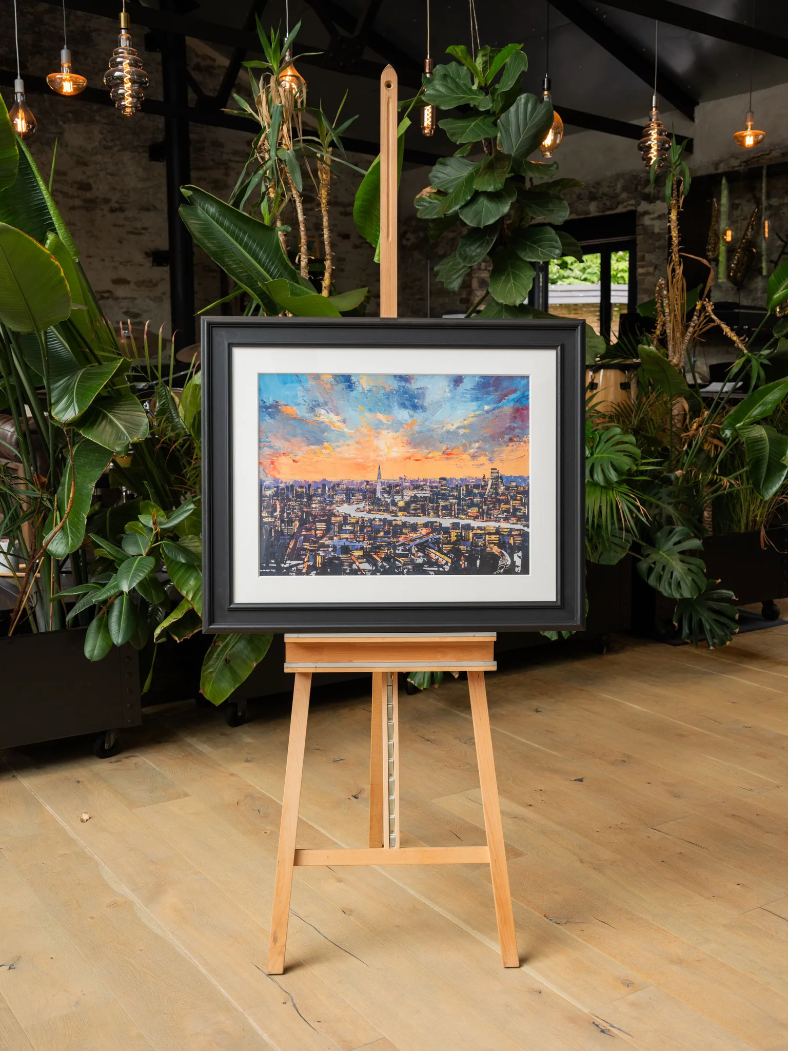 The City of Dreams, a limited edition print by Paul Kenton, in a black frame and white mount on a wooden easel among tropical plants, showing London at dusk under an orange and blue sky.