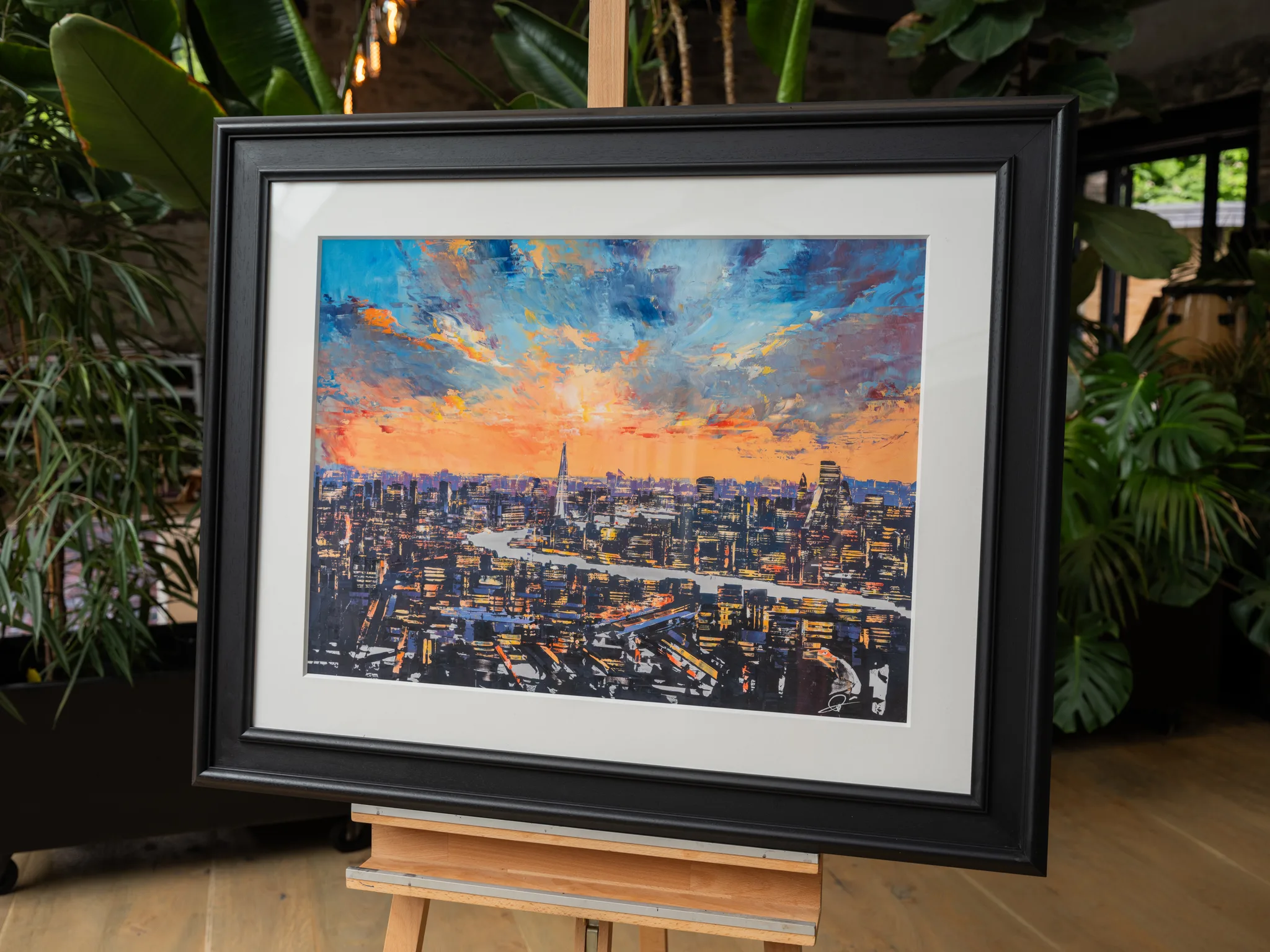 The City of Dreams, a limited edition print by Paul Kenton, shown in a black frame and white mount on a wooden easel among indoor plants. It depicts a London skyline at dusk, with a bright orange sunset under blue clouds above glowing city lights.