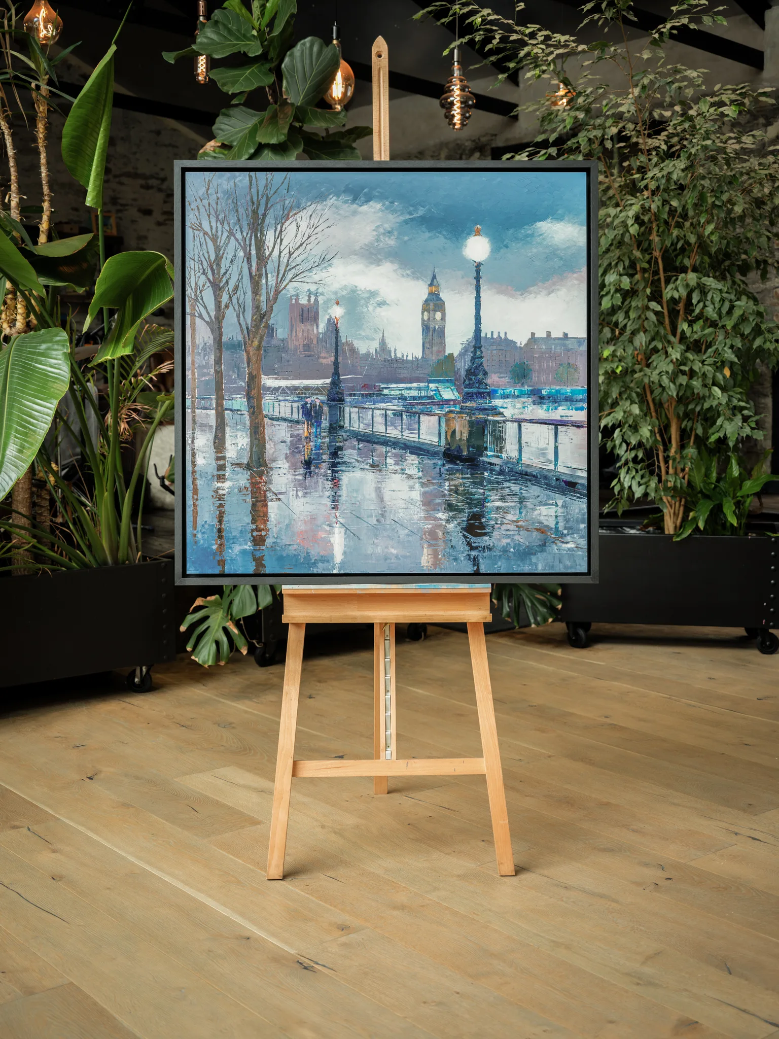 Thames Light, a limited edition print by Paul Kenton, shown in a dark frame on a wooden easel in a plant-filled room. It depicts a rain-wet London riverside walkway, with lamps, bare trees and Big Ben in blue and grey tones.