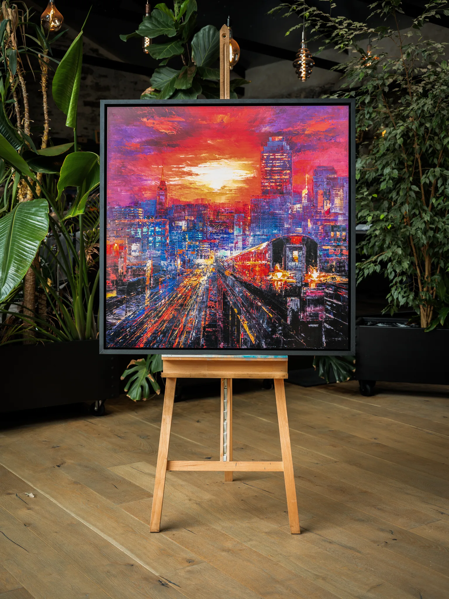 Urban Approach, a limited edition print by Paul Kenton, displayed on a wooden easel in a room of plants. Bold red and violet sunset over a glowing city, with a red train curving toward the viewer.