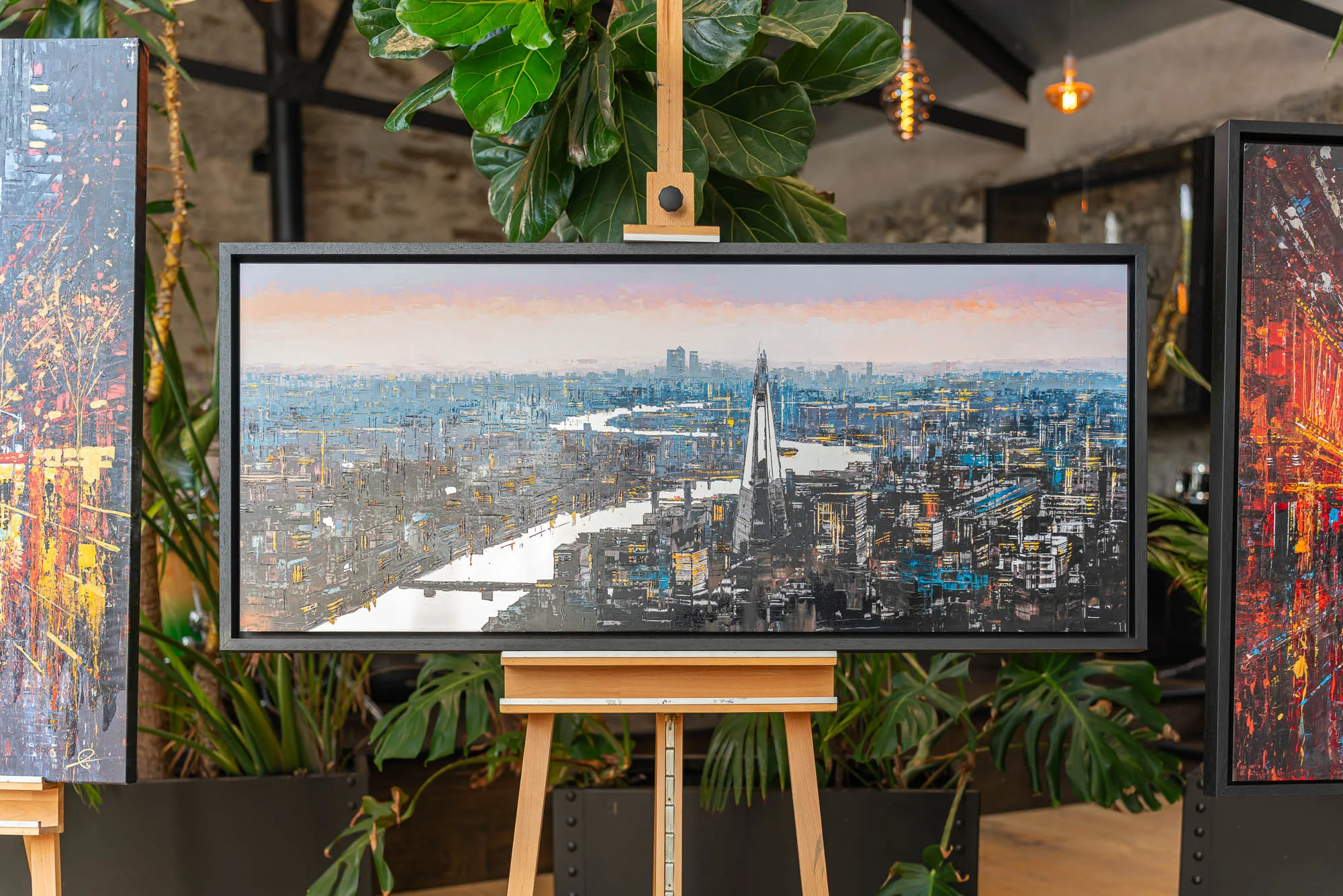 Vista Reflections, a limited edition embellished giclée print on aluminium by Paul Kenton, shown in a black frame on an easel among plants. It depicts a textured, twilight aerial view of London's Thames and skyline, with a tall glass tower in the centre.
