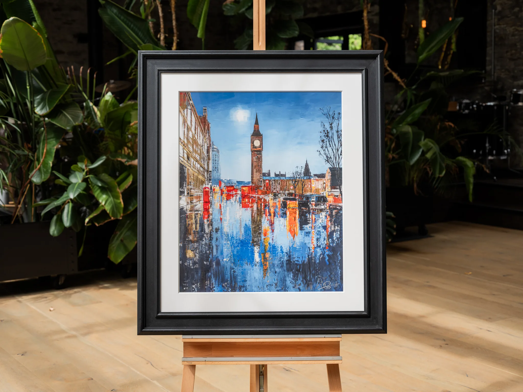 Westminster Illuminates, a limited edition print by Paul Kenton, in a black frame and white mount on a wooden easel among indoor plants. It shows Big Ben under a pale blue sky, with red and orange reflections rippling across blue water.