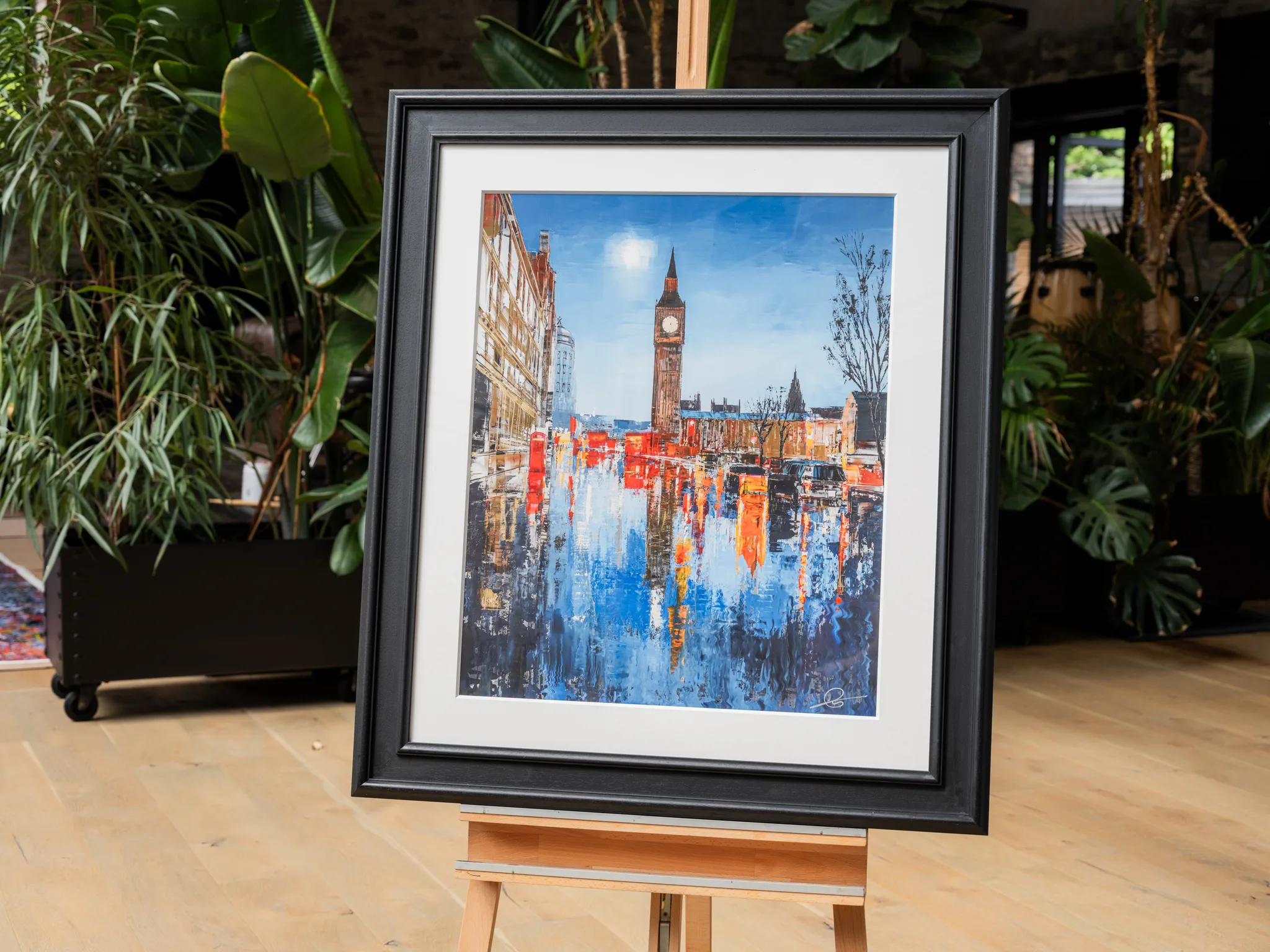 Westminster Illuminates, a limited edition print by Paul Kenton, shown in a black frame and white mount on a wooden easel among indoor plants. Big Ben rises above a street of warm orange and blue reflections.