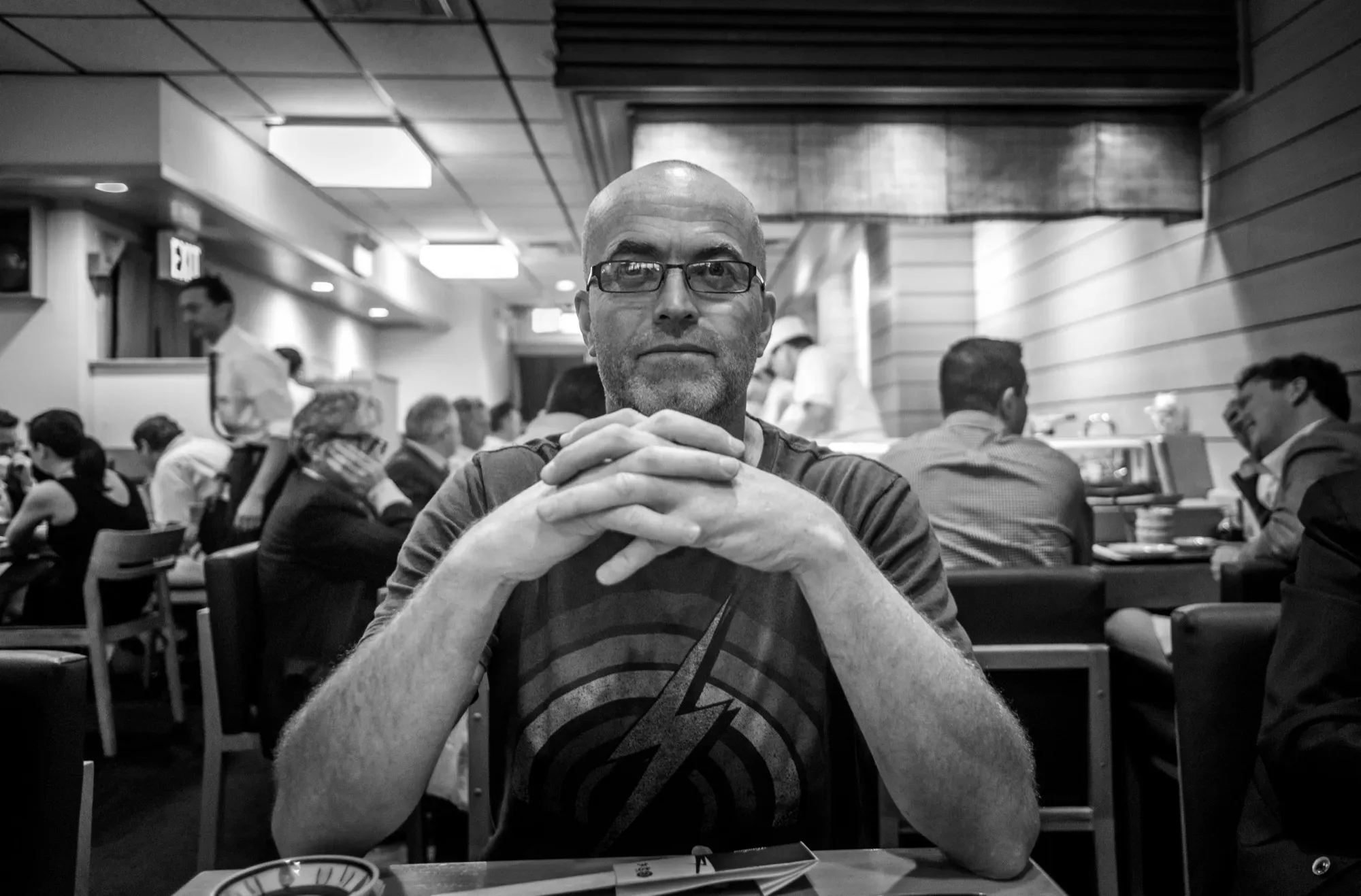 Black-and-white photo of Paul Kenton sitting at a table in a busy New York restaurant, hands clasped